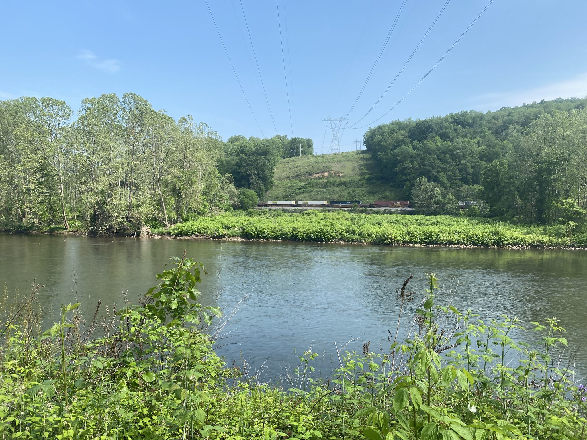 A wide river with green banks, distant train, and power lines under a sunny sky. - Roundbottom Trekker Campground – The Mailbox formation right beside the trail loop from West Newton