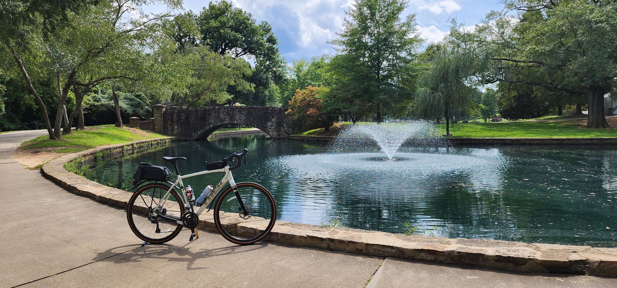A bicycle parked by a lake with a fountain and a stone arch bridge. - Westfield Rd. at Brandywine – Fountain at Freedom Park loop from Queens University
