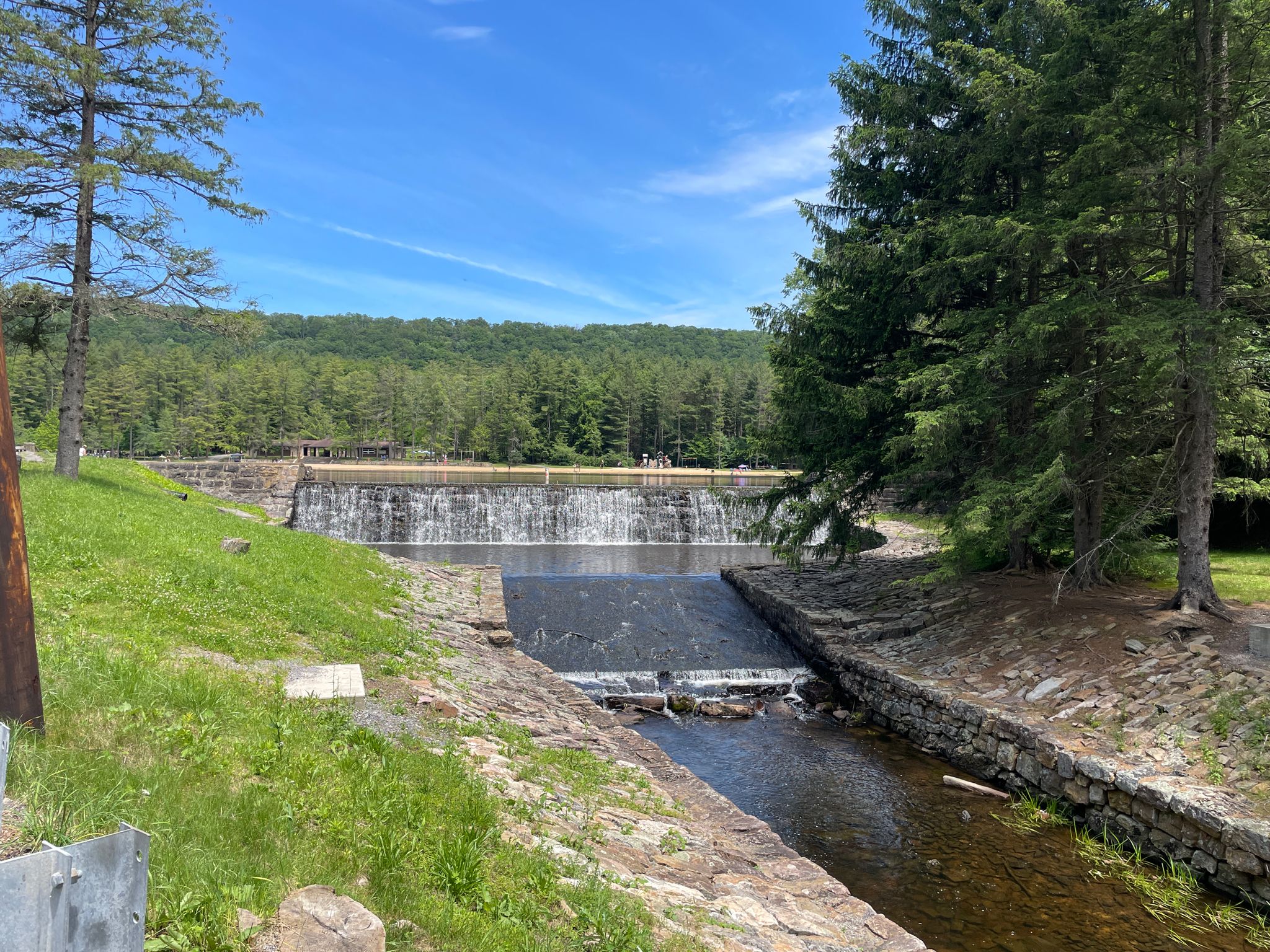 Sunny day at a dam with water flowing, surrounded by green trees and distant people. - Bake Oven and Mid-State Trail Loop