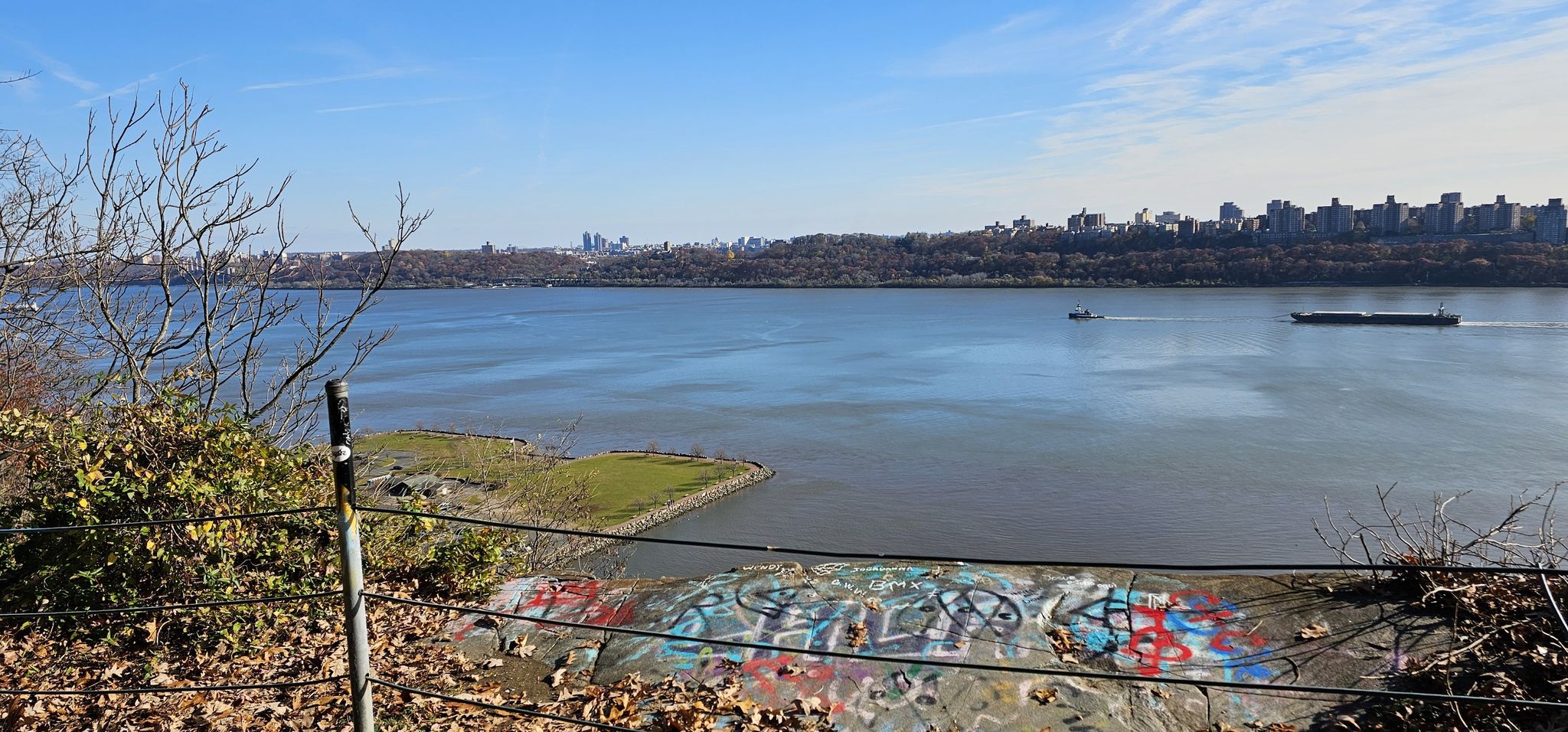 River view from above with graffiti, distant city, and two boats on a bright day. - Long Path and Shore Trail Loop