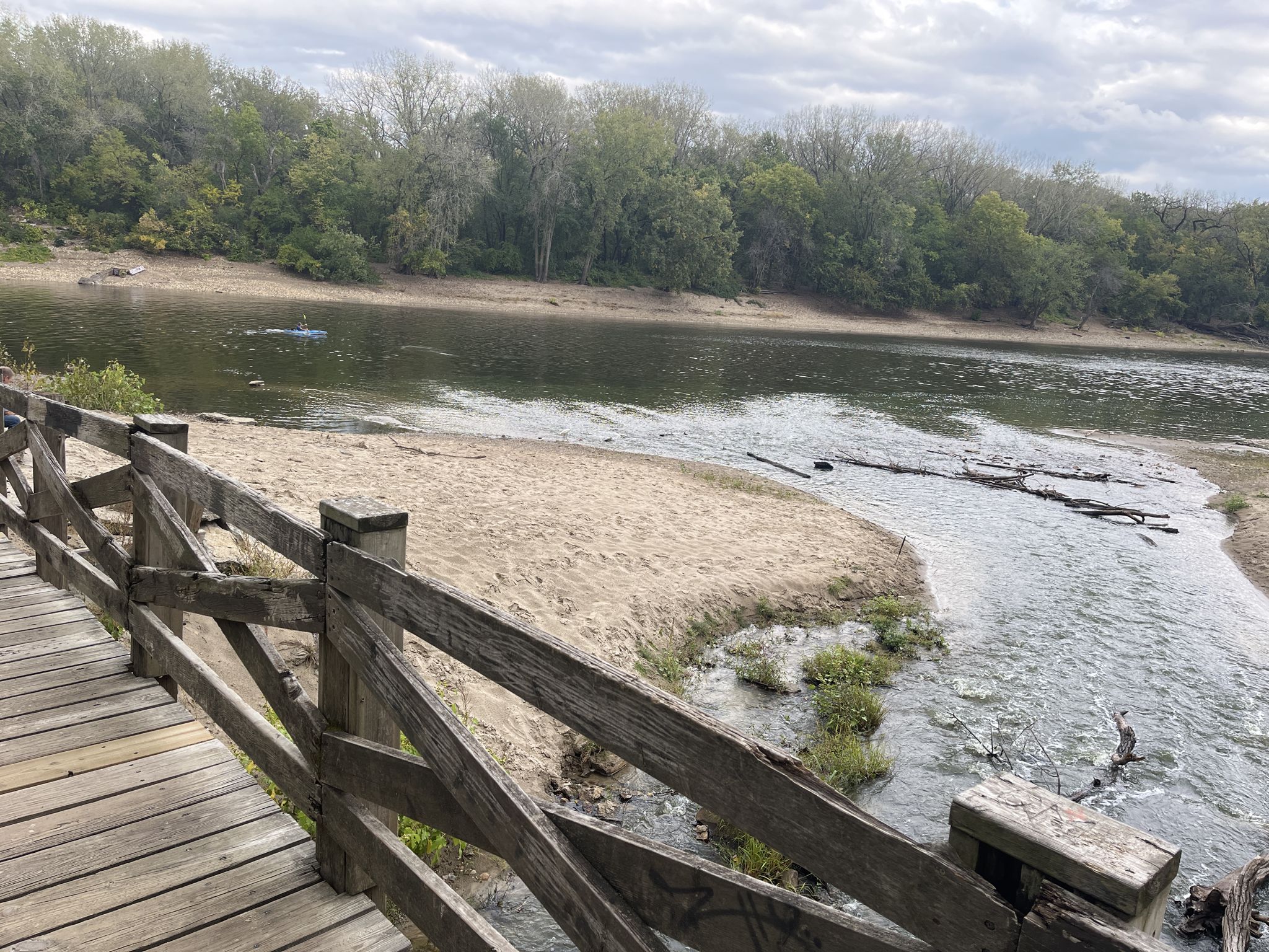 Wooden bridge railing overlooking a river, sandy bank, and green forest. - Minnehaha Regional Park loop