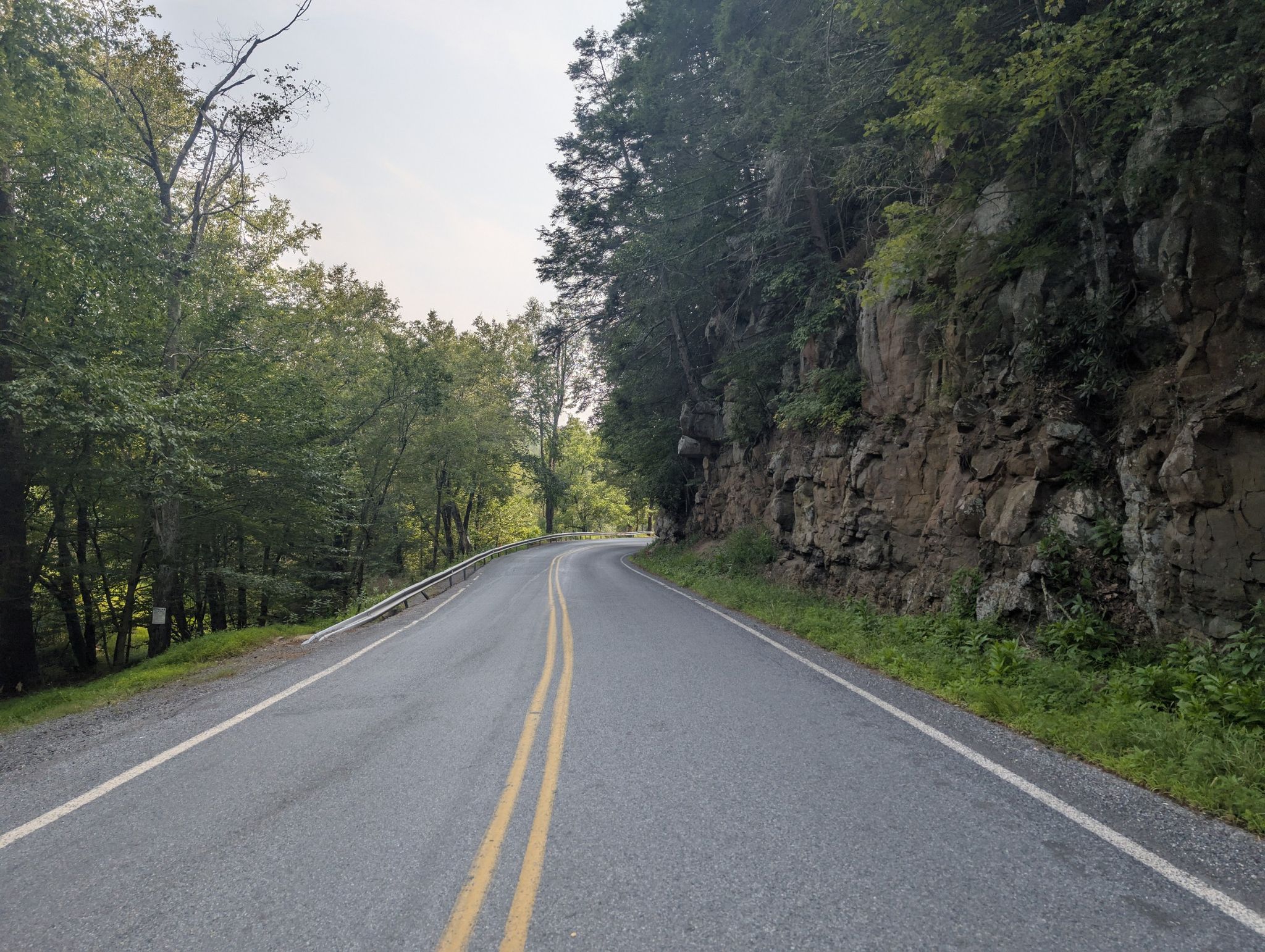 Empty paved road curves alongside a rocky cliff and dense forest. - White Haven – D&L Trail loop from Maple Tract Preserve