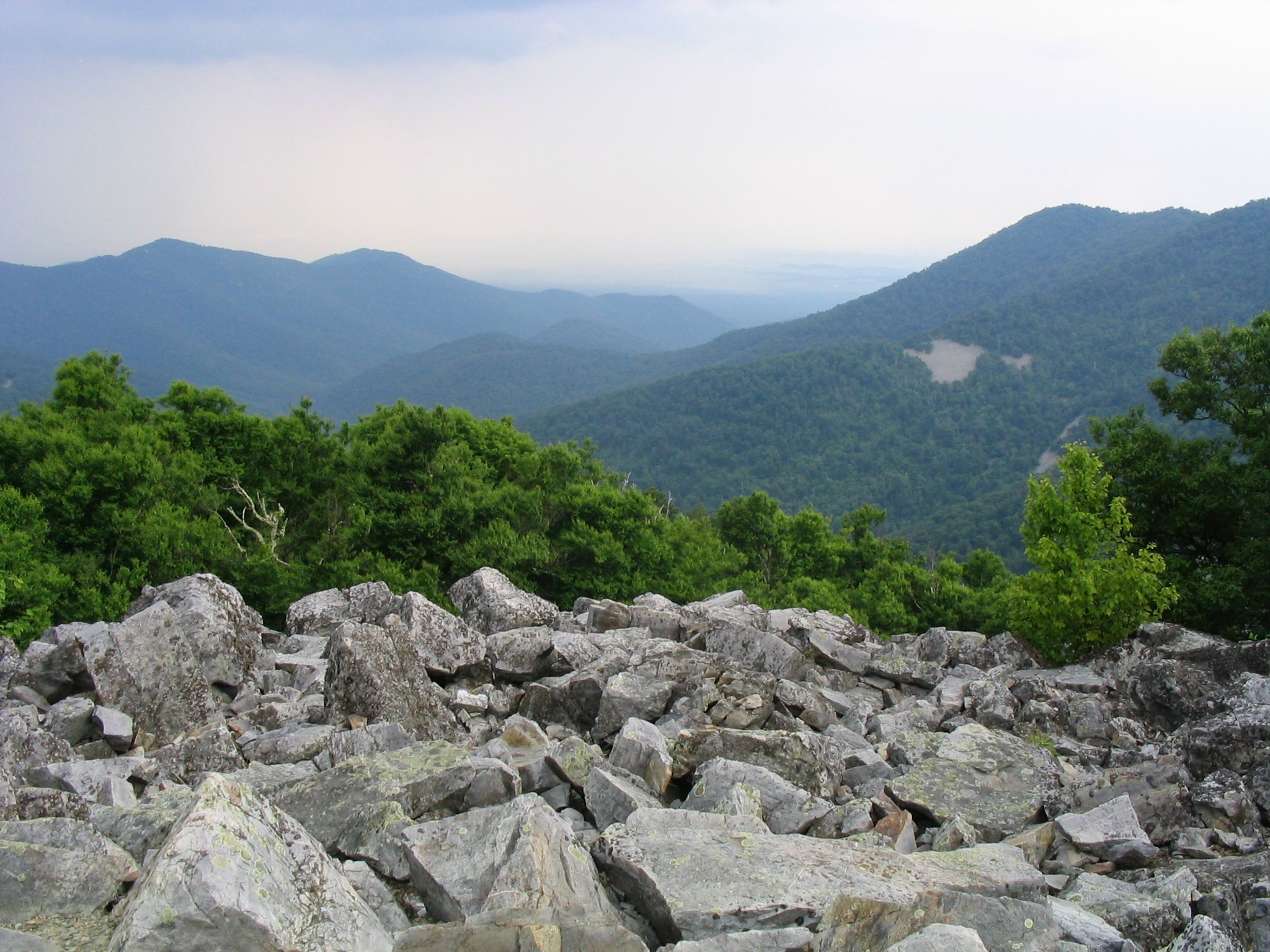 Overcast view of green mountains and valleys with a foreground of large gray rocks. - Blackrock Summit loop