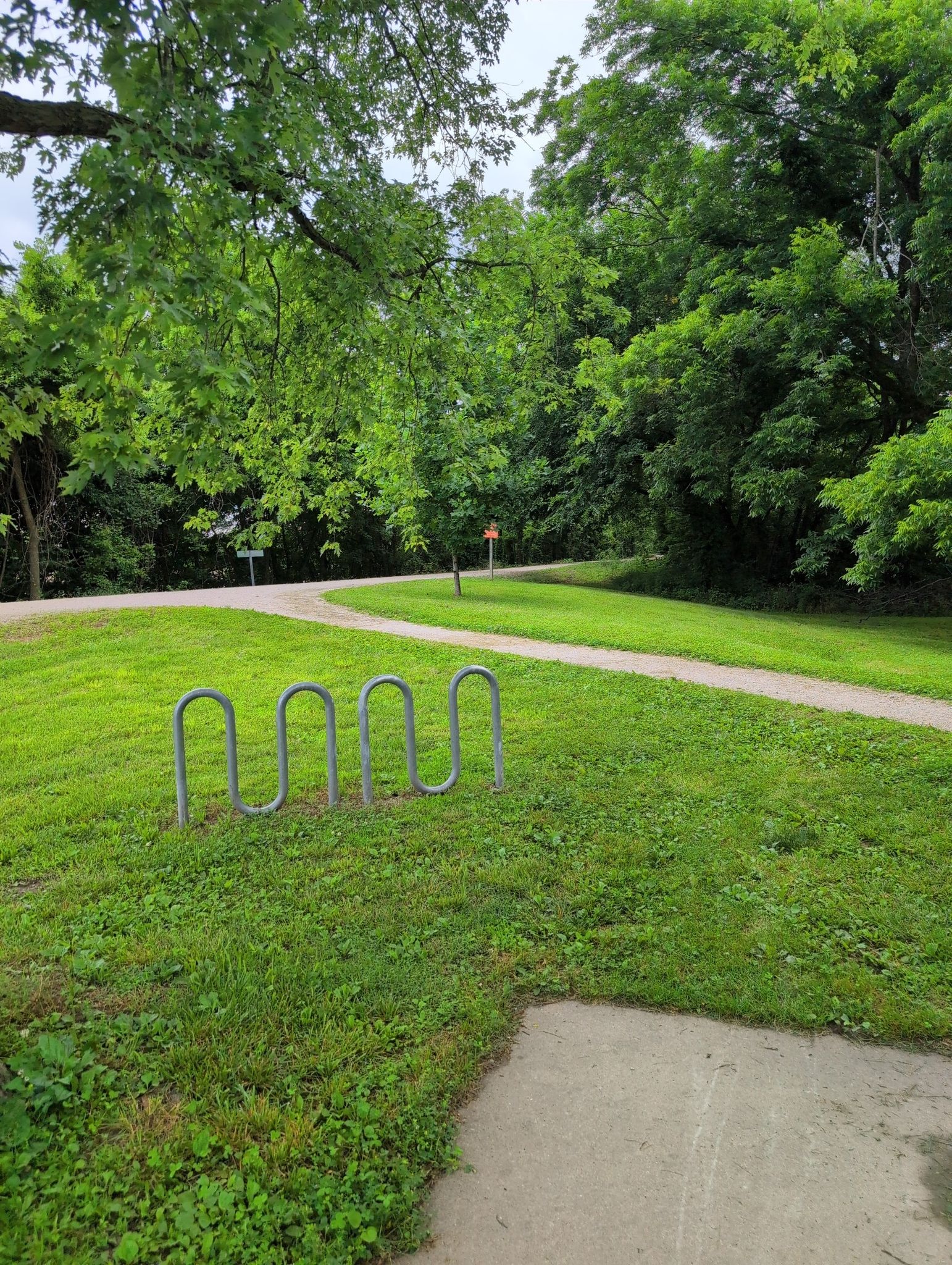 A metal bike rack sits on green grass next to a winding paved path. - Katy Trail: Dutzow - Marthasville