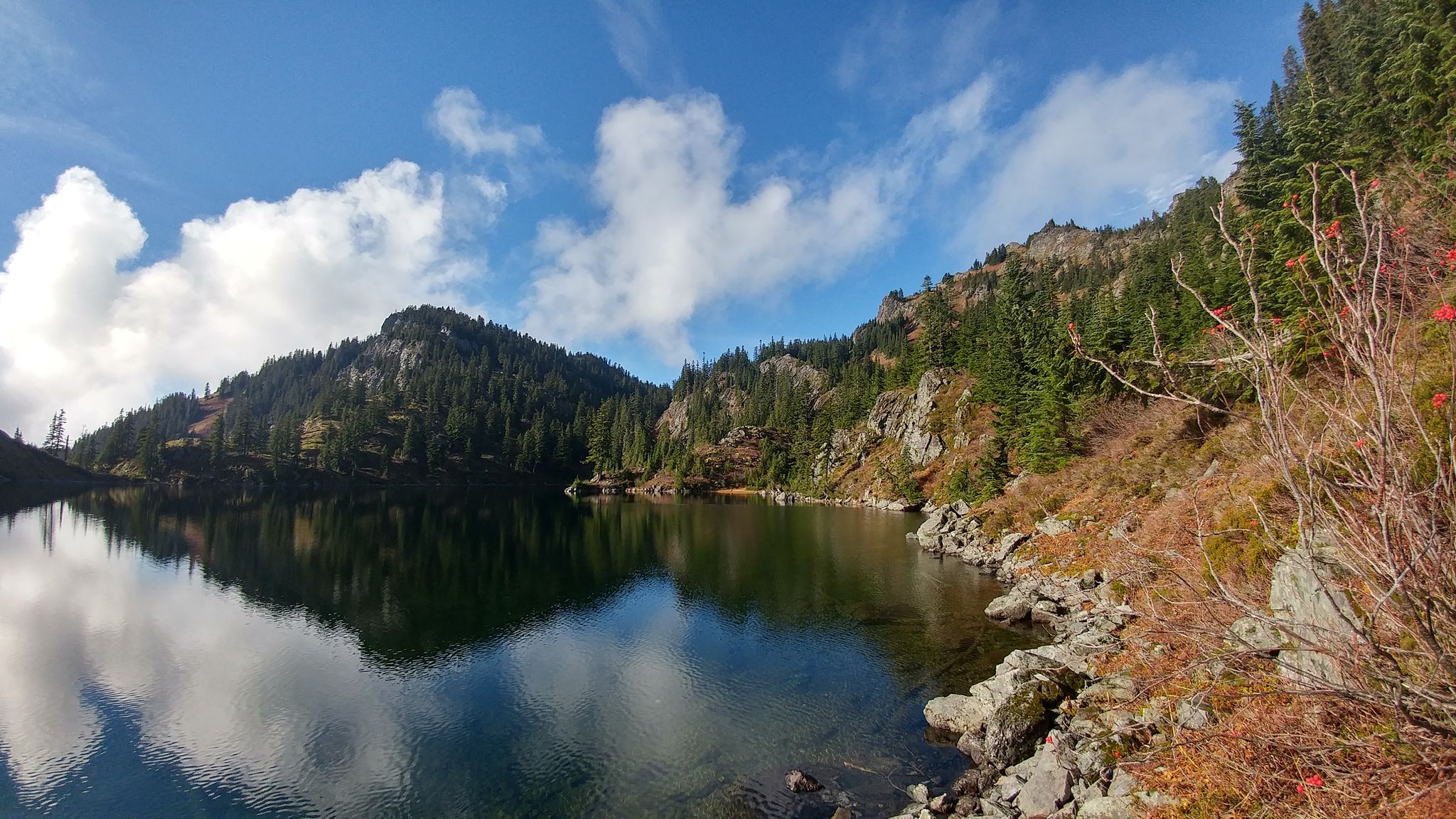 Calm mountain lake reflecting blue sky, surrounded by rocky, forested slopes. - Lake Lillian Trail