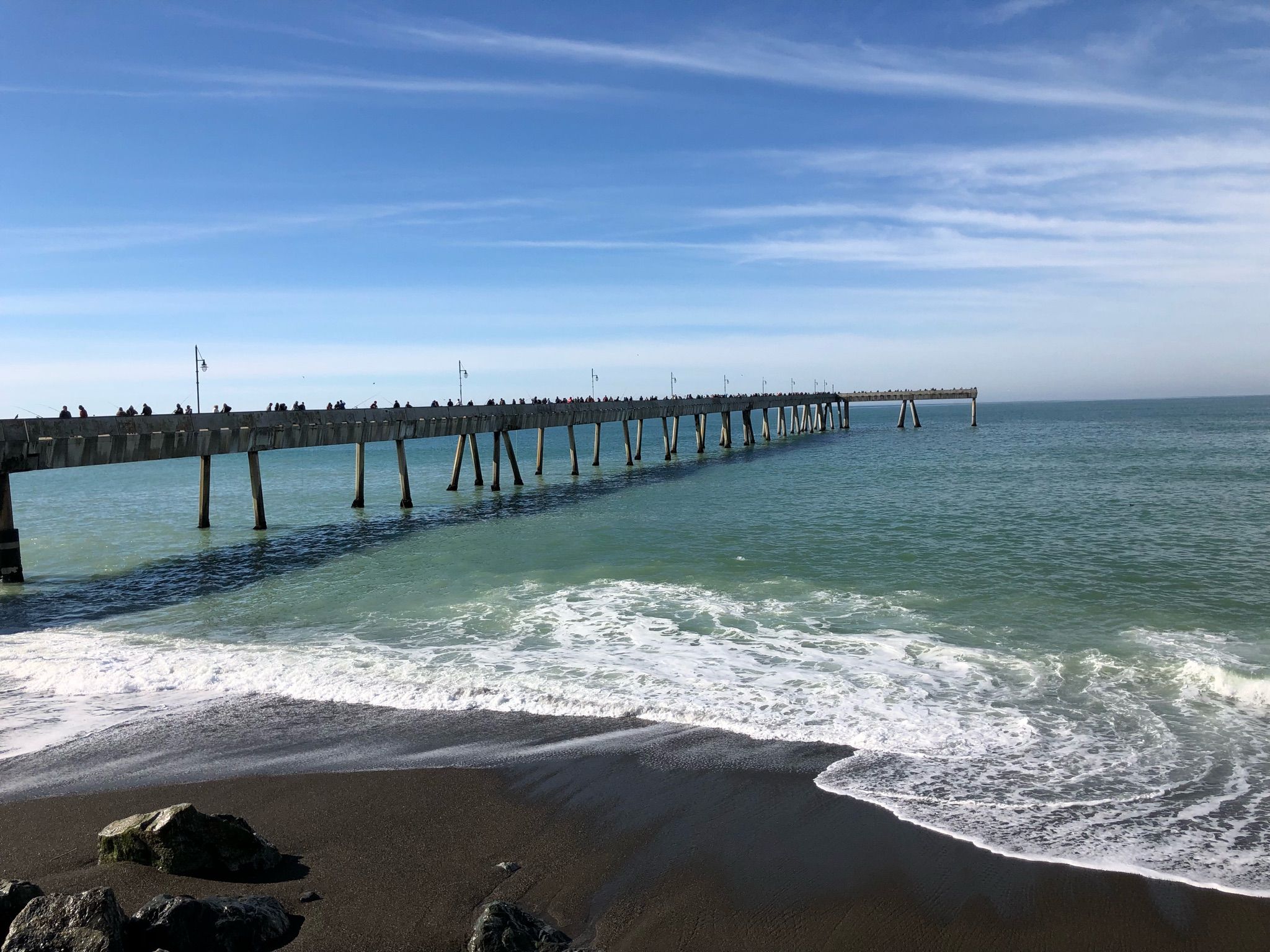 Long pier extends over the blue ocean with people and crashing waves. - Sharp Park Beach – Rockaway Beach loop from Milagra Ridge