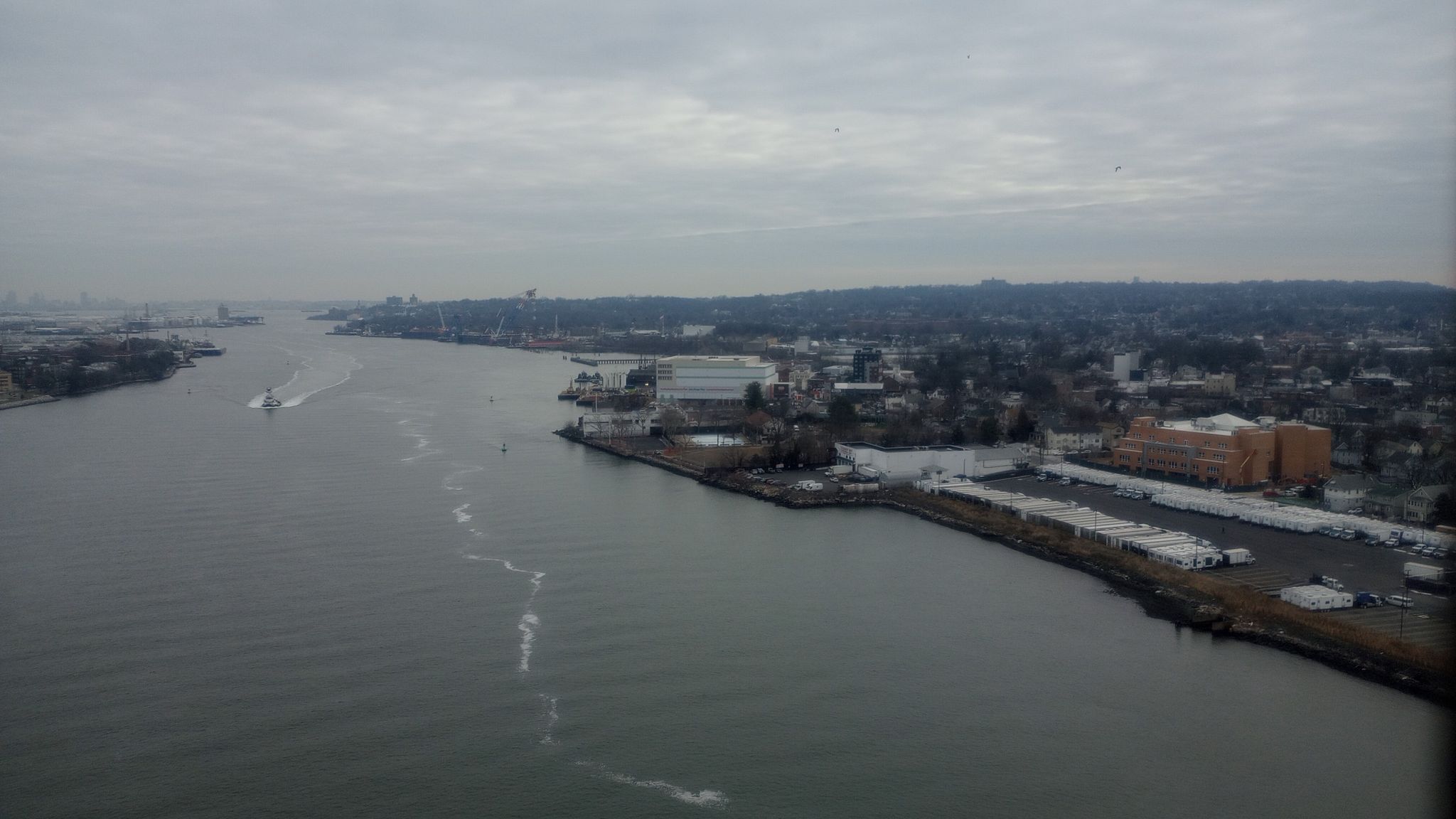 Wide river with a boat, urban banks, and industrial buildings under a gray sky. - Stephen R. Gregg Park – Bayonne Bridge loop from Danforth Avenue