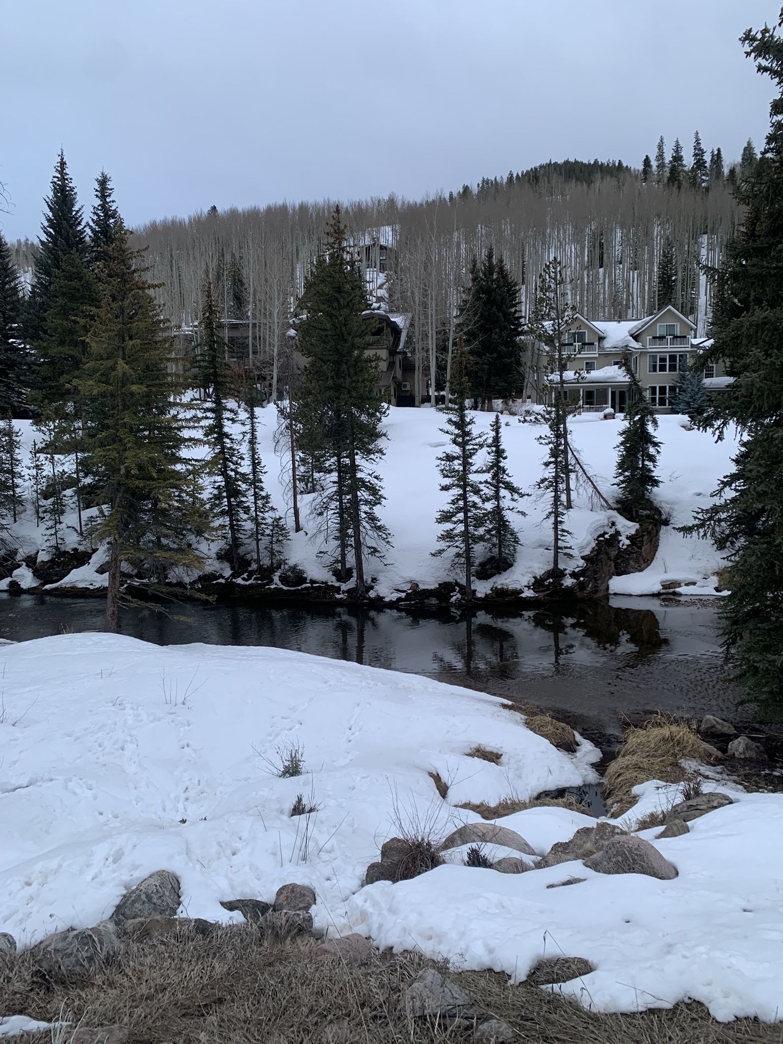 Snowy riverbank with dark water, evergreen trees, and a large house under an overcast sky. - Gore Valley Trail