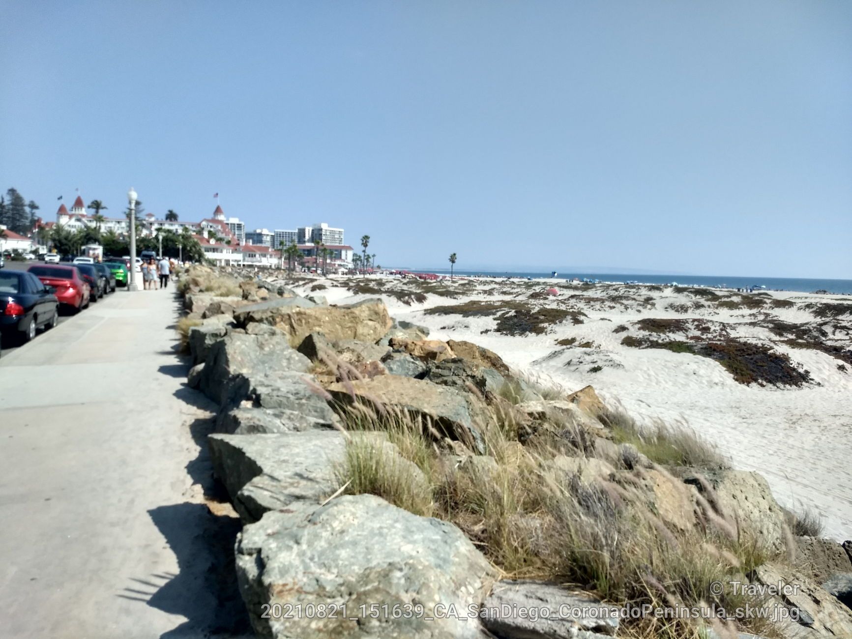 Sidewalk, rocky barrier, sandy beach, ocean, and distant buildings under bright sun. - San Diego Bridge – Coronado Ferry Landing loop from Coronado