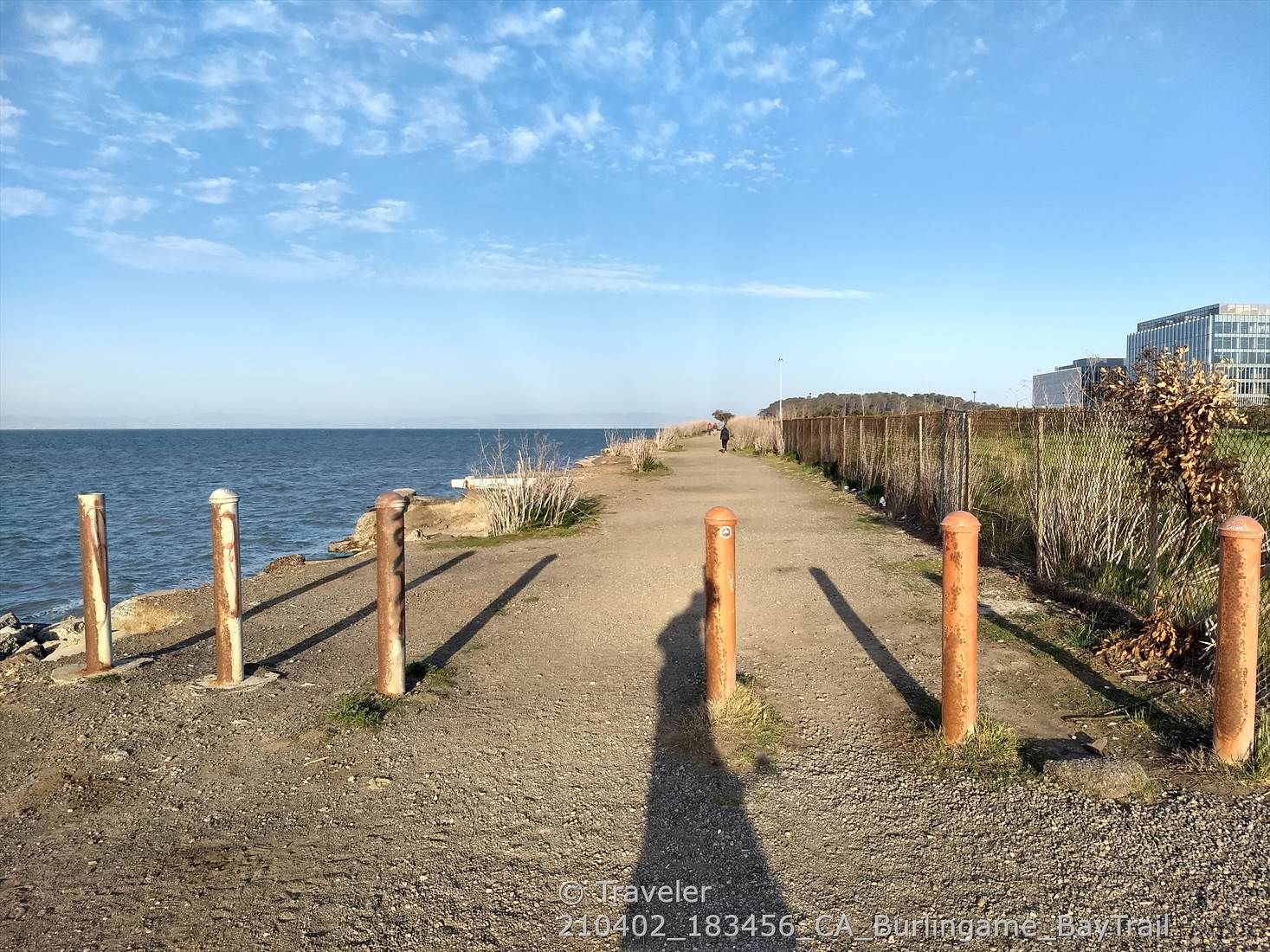 Dirt path by water with wooden posts, person walking, under a bright sky. - San Francisco Bay Trail: Burlingame to Redwood Shores
