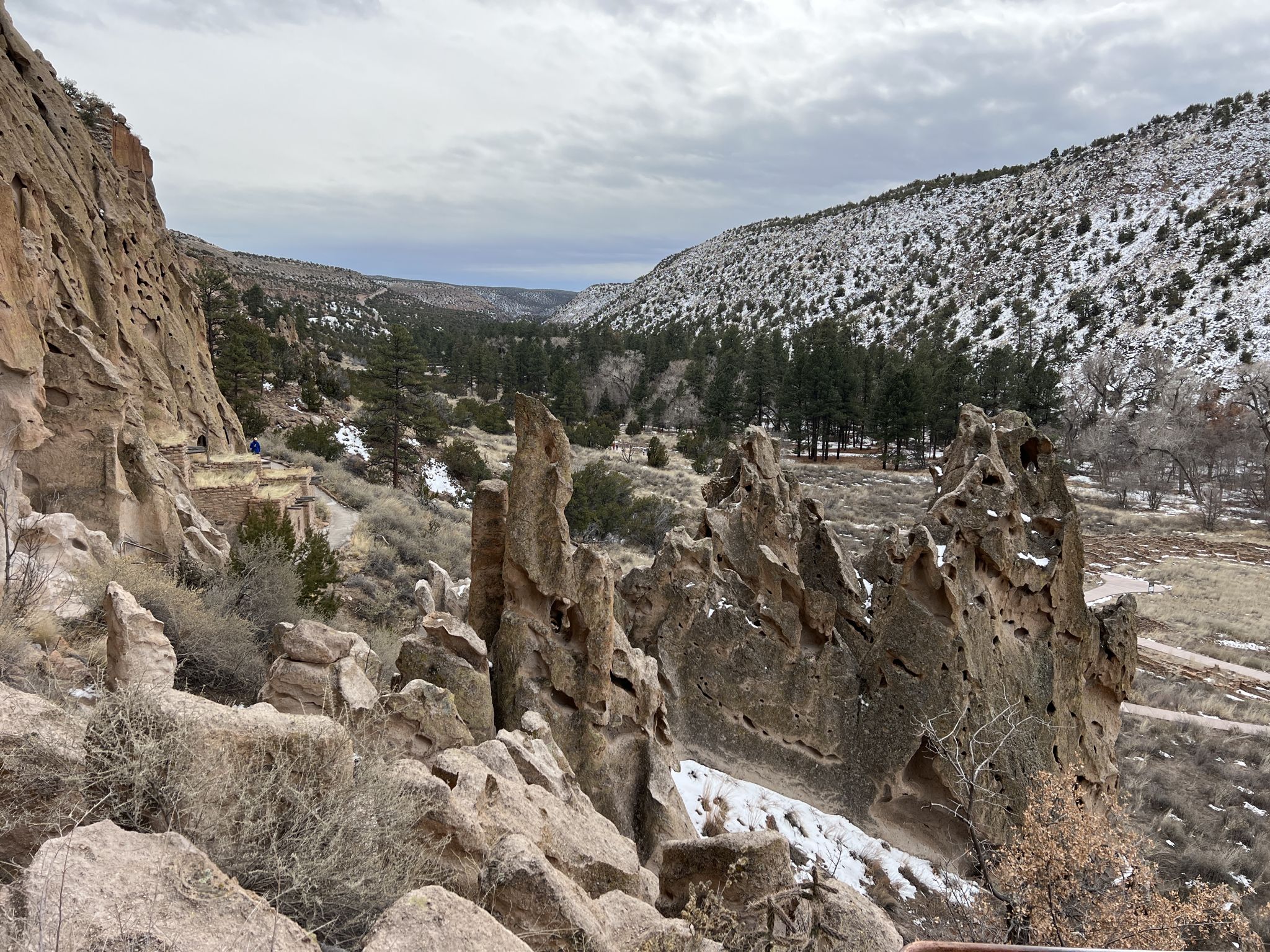 A wide valley with unique rock formations and snow-dusted hills under a cloudy sky. - Pueblo Loop Trail