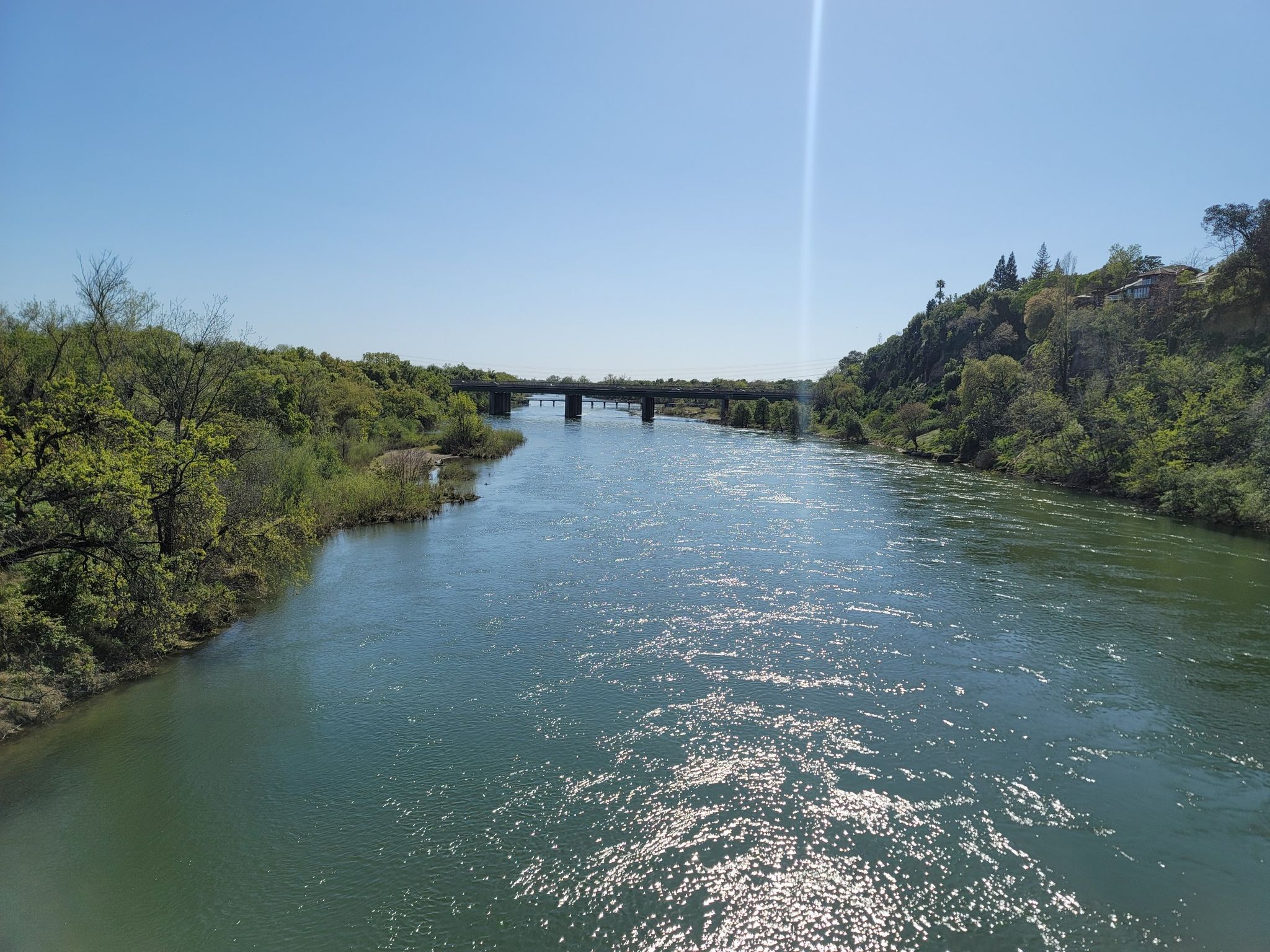A wide river flows under a bright blue sky with a distant bridge. - Fair Oaks Bridge loop from Gold River