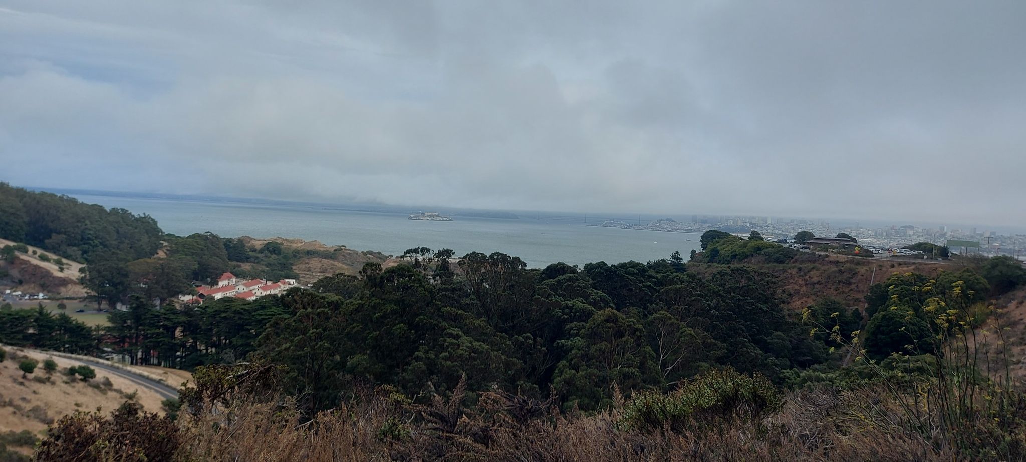 Overcast view of a bay surrounded by green hills and distant buildings. - Sausalito Yacht Harbor – Downtown Sausalito loop from Sausalito