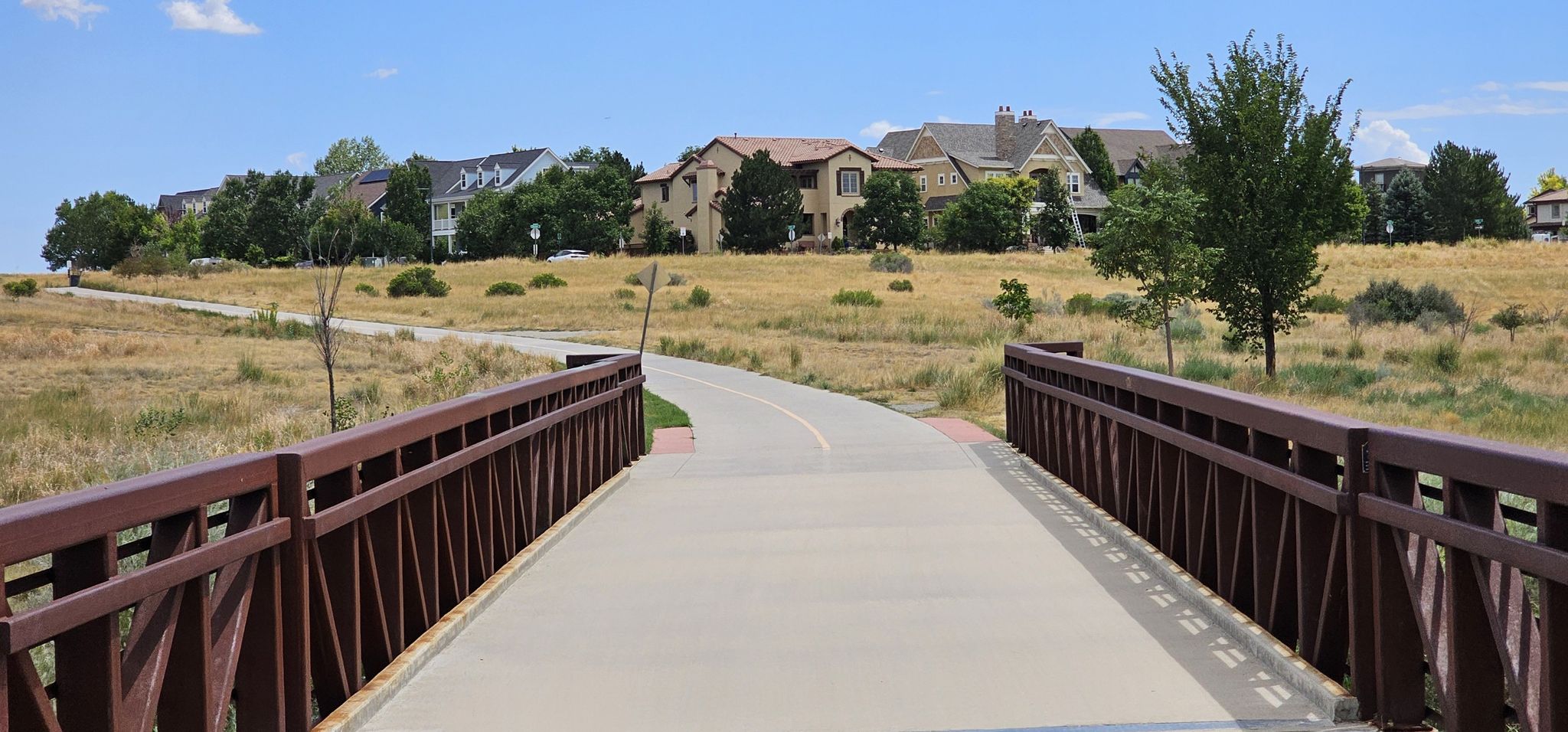Concrete bridge and paved path through dry grass, with modern houses in the background. - Westerly Creek Trail