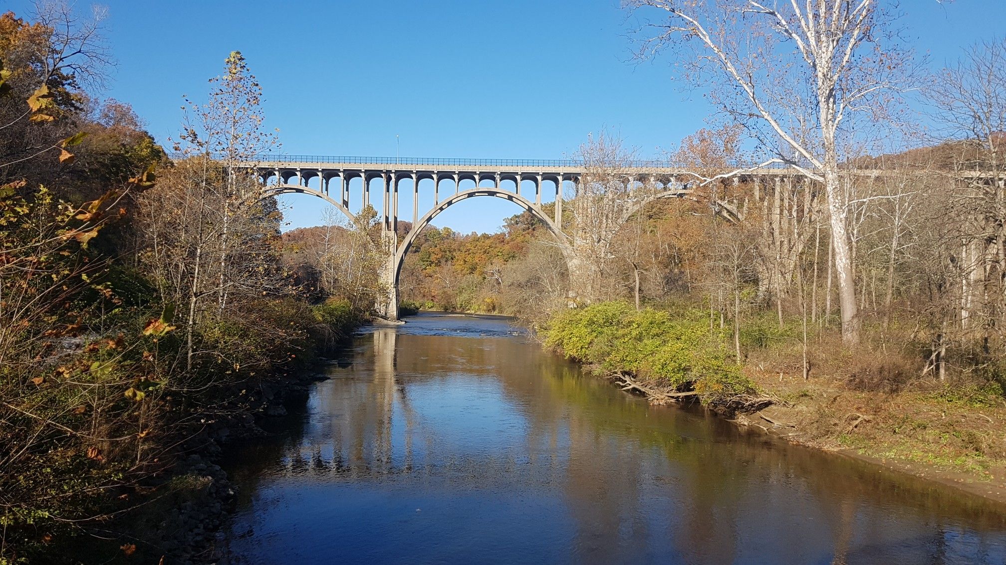 A large arched bridge spans a river, surrounded by autumn trees under a clear sky. - Brecksville-Northfield Bridge – Deer Lick Cave loop from Brecksville