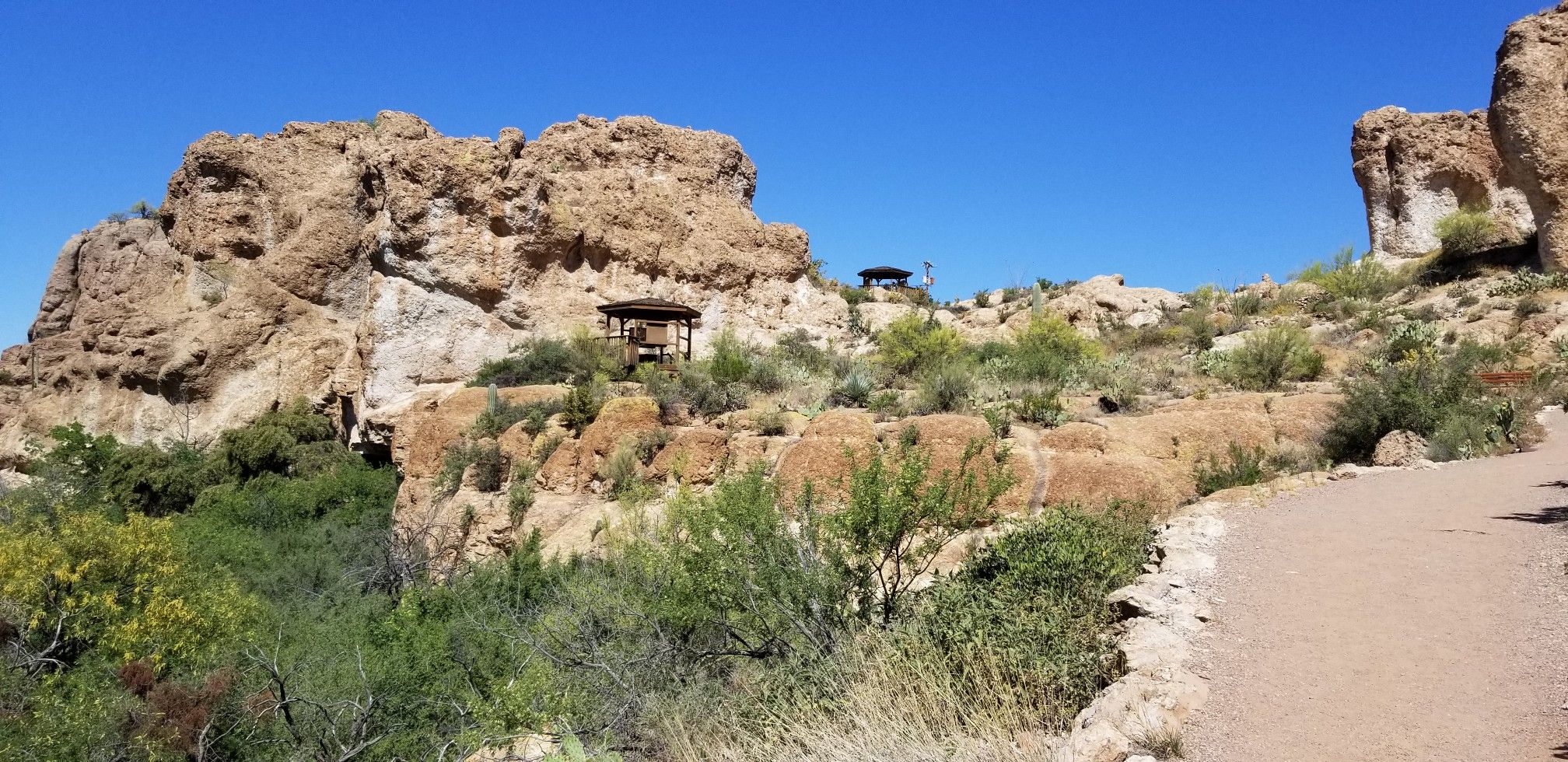 A winding dirt path through rocky desert hills with sparse green shrubs and shelters. - Main Trail