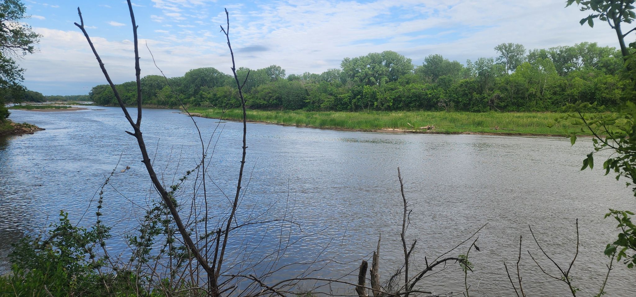Wide river flows past green banks under a bright, cloudy sky. - Manhattan Linear Trail
