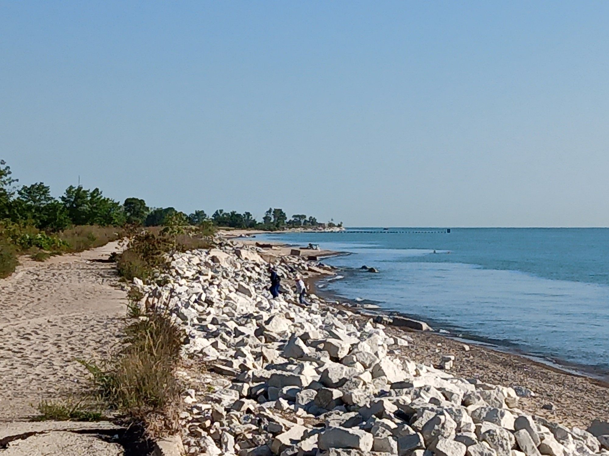 Sunny day, dirt path next to rocky shore and calm blue water with a person. - Illinois Beach Trail