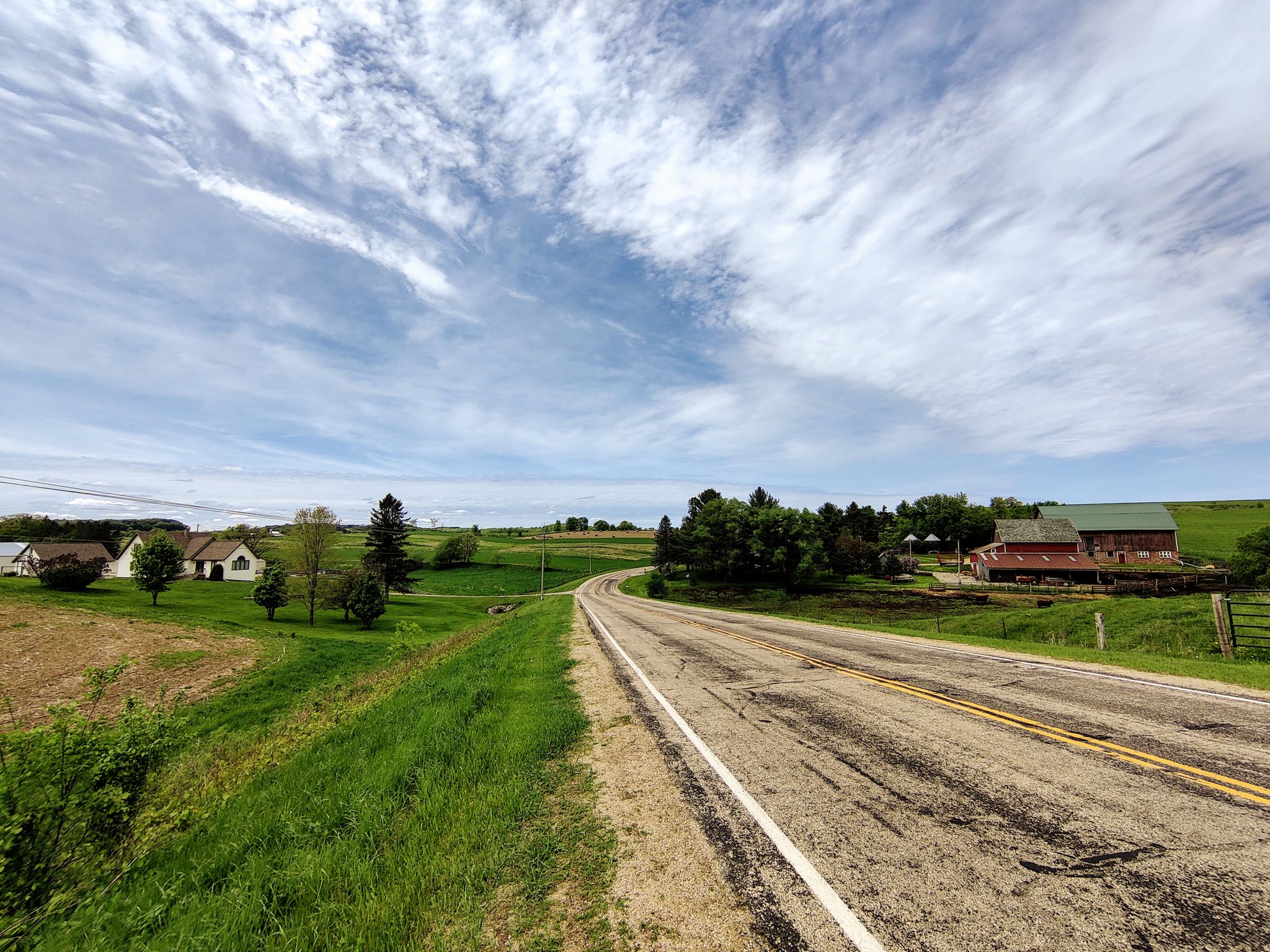 A winding road through green rolling hills with distant farmhouses under a cloudy sky. - La Valle and Back loop