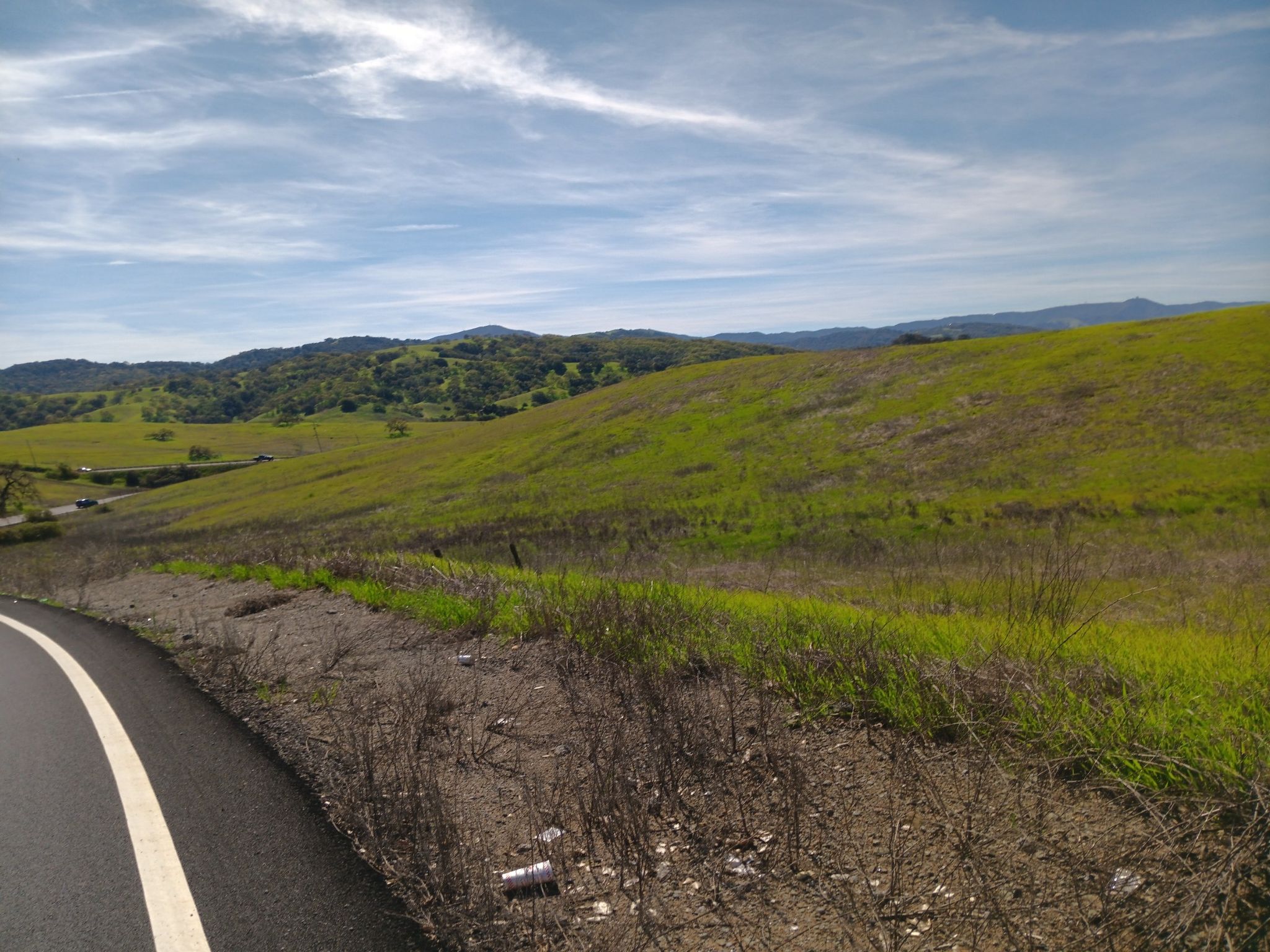 Paved road next to a grassy hillside with distant green mountains under a blue sky. - Bailey Road