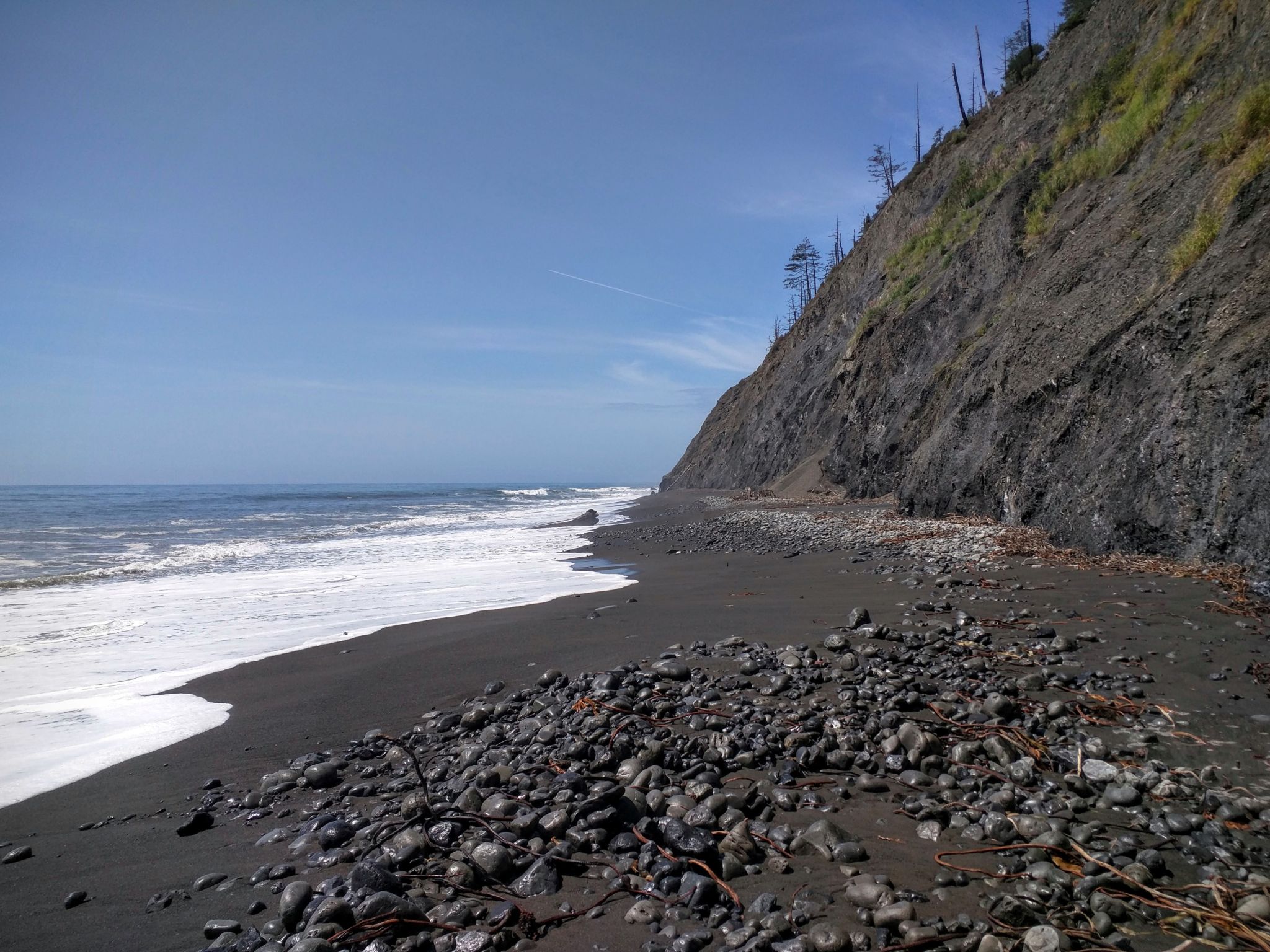Sunny dark rocky beach with white waves crashing against a steep cliff. - Hammocker's Gulch 🌊🚰 – Sliding jungle cliff loop from Shubrick Peak