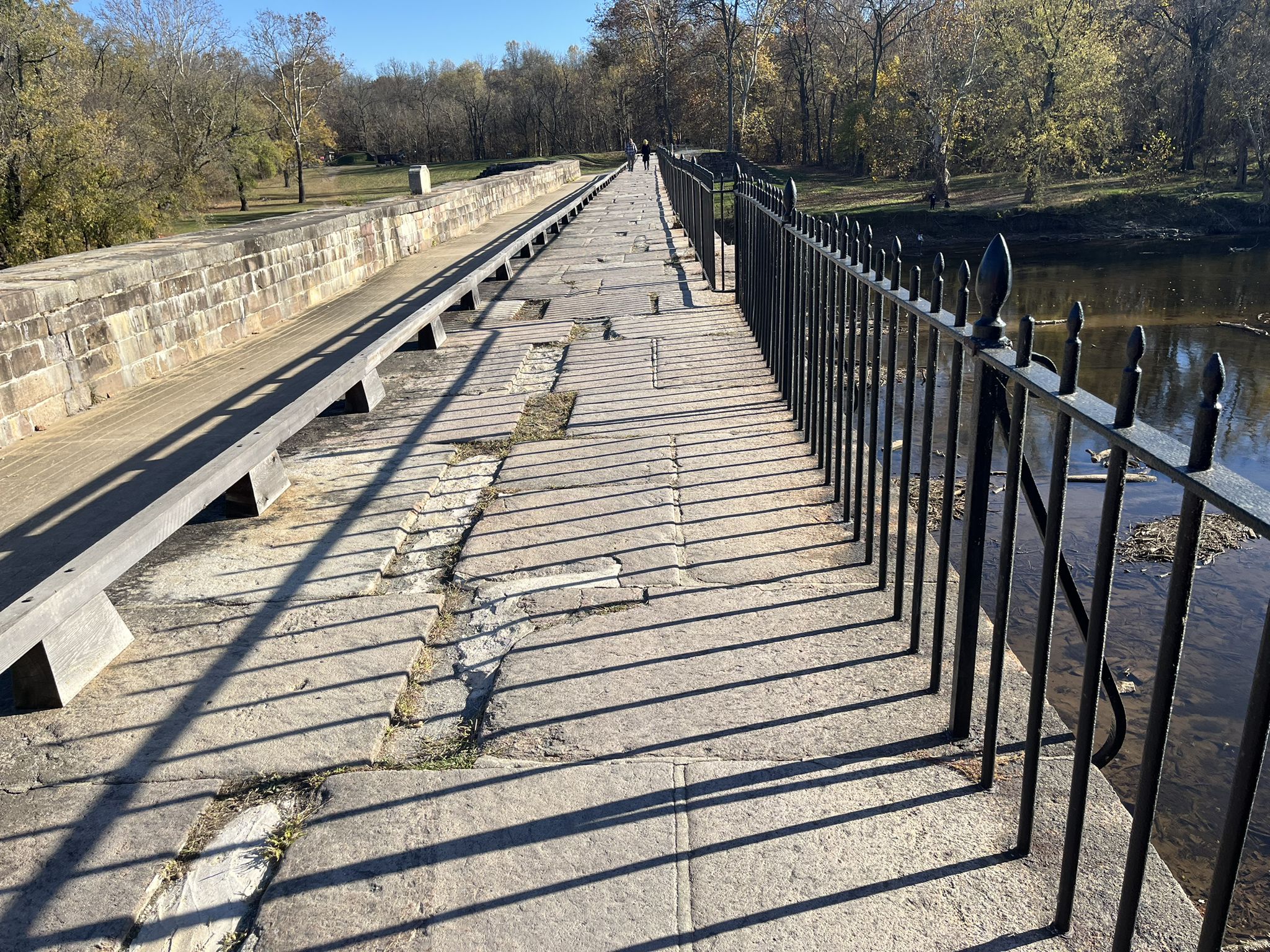 Stone path on a bridge with a black railing, long shadows, and autumn trees. - Chesapeake and Ohio Canal Trail