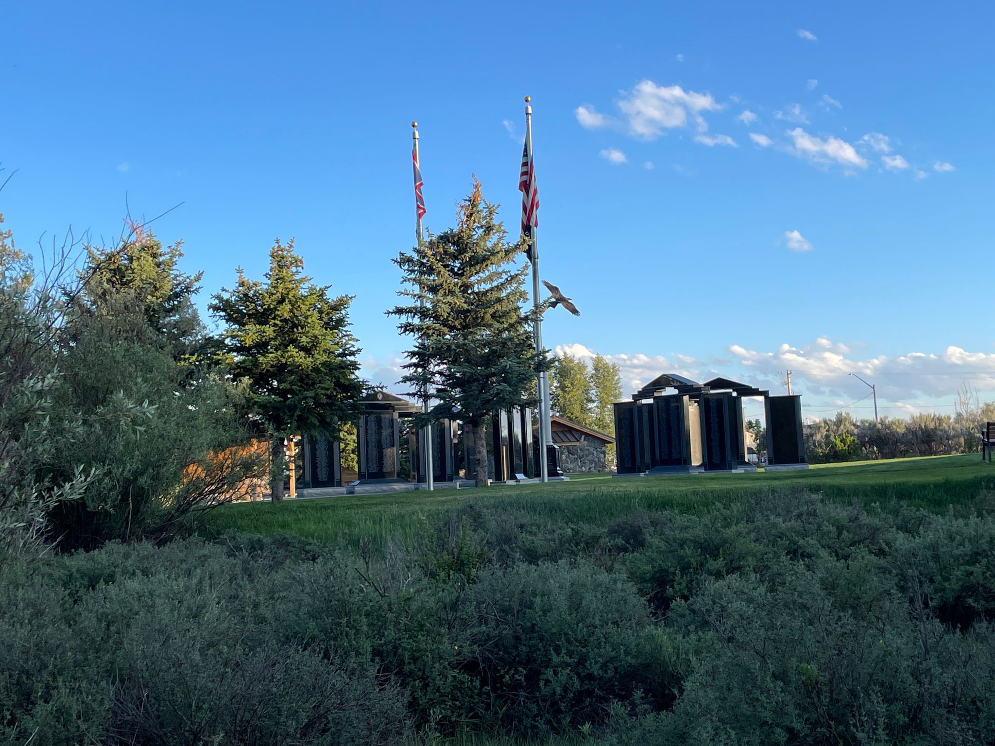 A memorial with flags, evergreen trees, and dark structures under a blue sky. - Boyd Skinner Trail