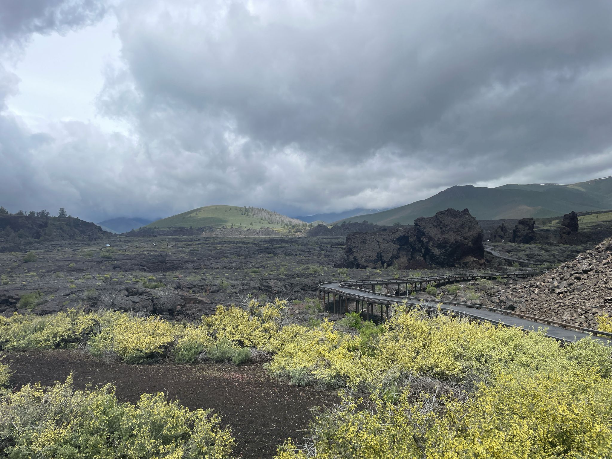 Winding boardwalk through dark volcanic rock and green shrubs under cloudy sky. - North Crater Flow Trail