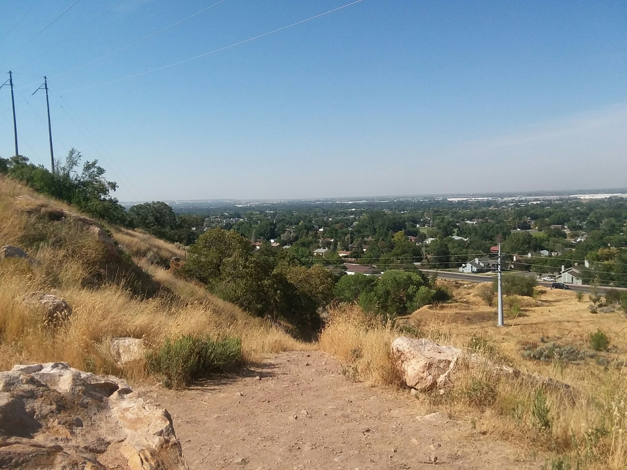 Dirt path descends a dry, grassy hill overlooking a distant town under clear skies. - Ogden Nature Center North loop from Ogden Nature Center North