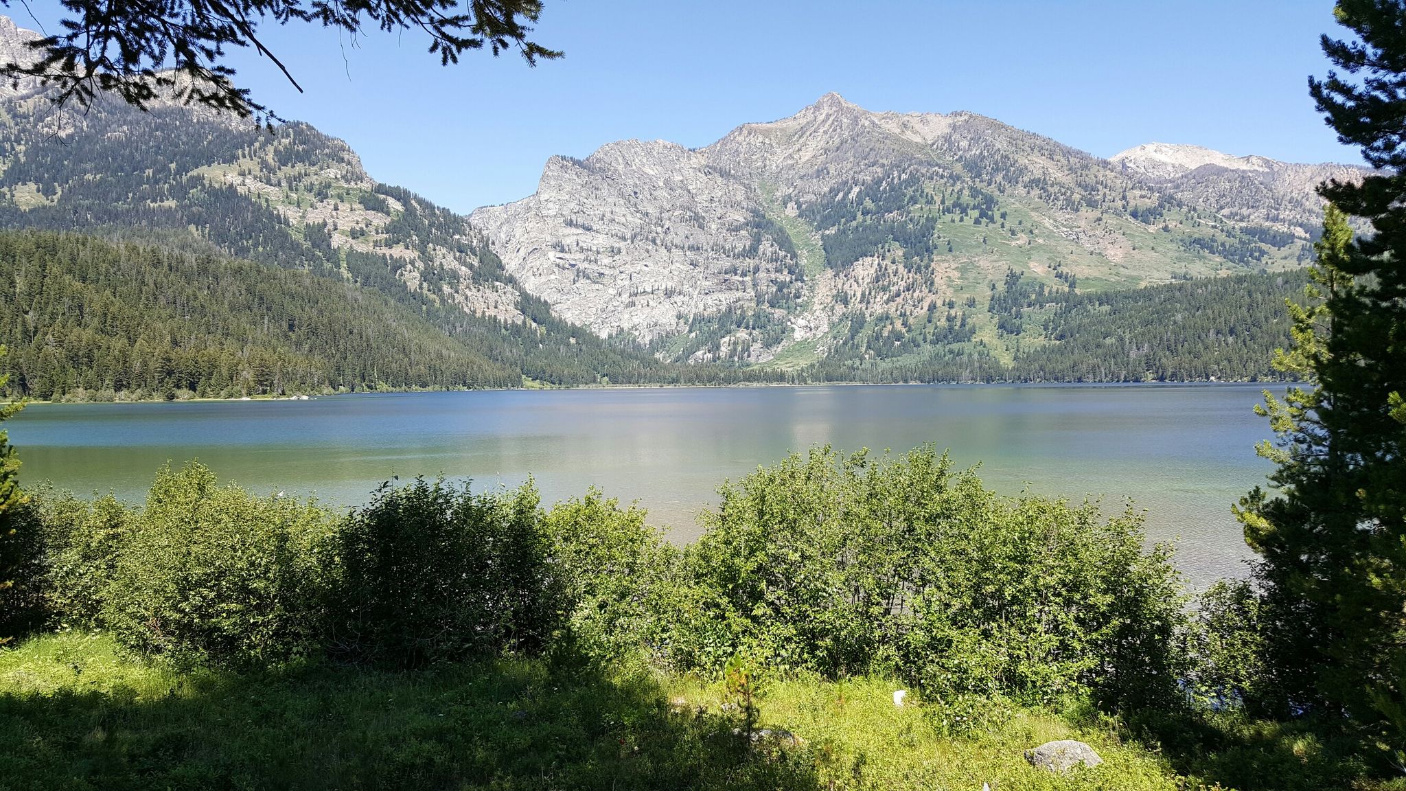 A serene lake surrounded by lush green mountains under a bright sky. - Lake Creek Trail