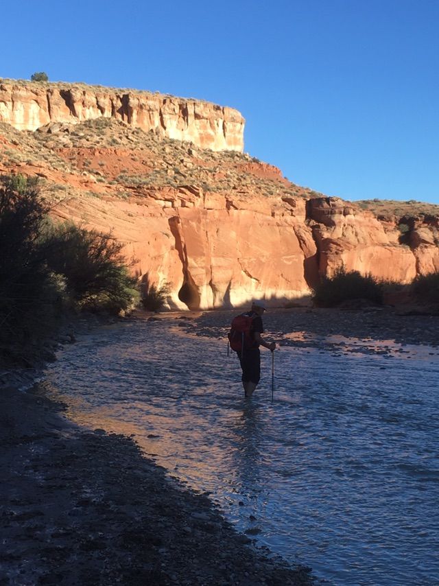 A person with a backpack wades through a shallow river in a canyon. - White House / Paria River Trail