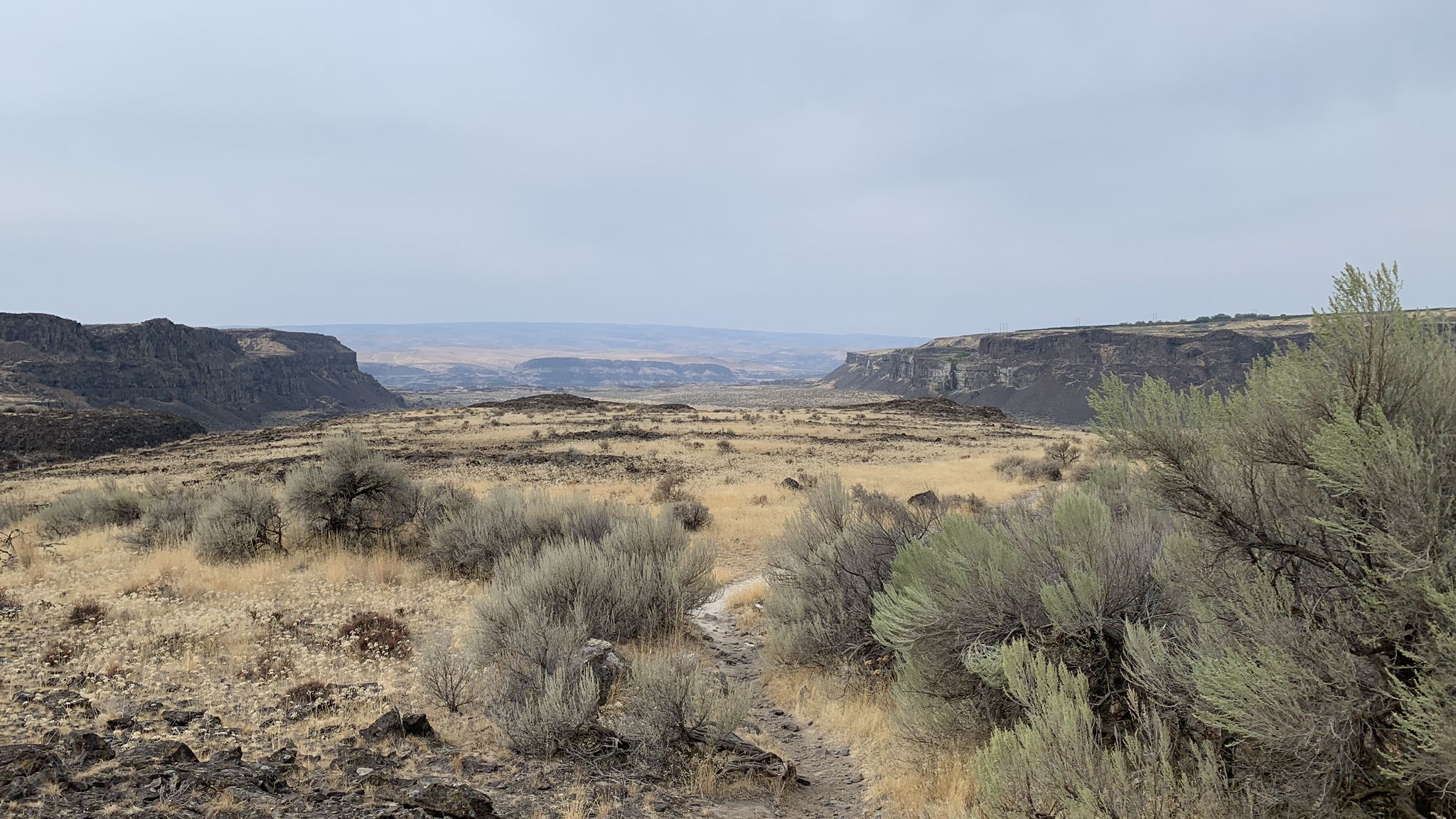 A wide, arid canyon with dry brush and distant hills under a cloudy sky. - Dusty Lake Trail