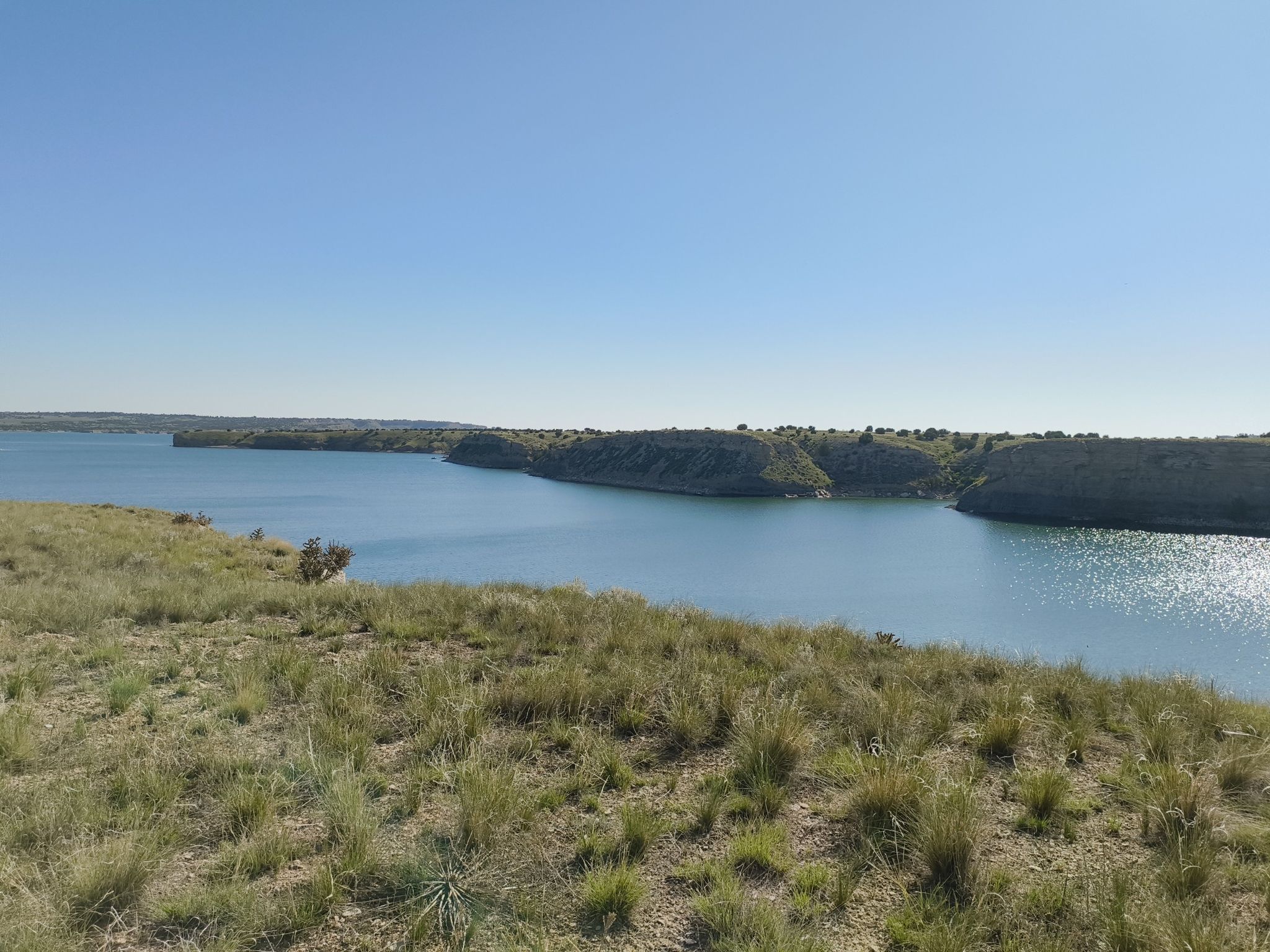 Expansive blue lake surrounded by green and brown grassy hills under clear sky. - Voodoo Loop