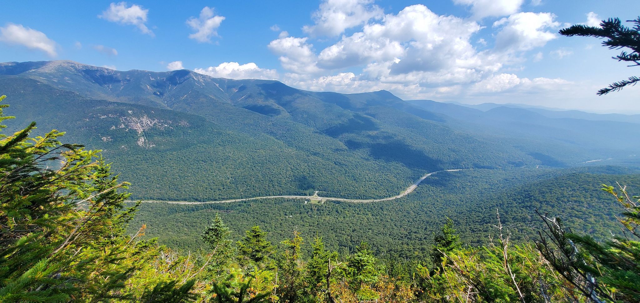 Panoramic view of green mountains, a valley road, and a bright sky. - North Kinsman, South Peak and Cannon Balls Loop Trail