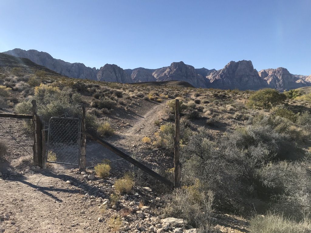 Rustic gate on a dirt path leading to arid mountains under a clear sky. - Shattered Glass and Landmine Loop