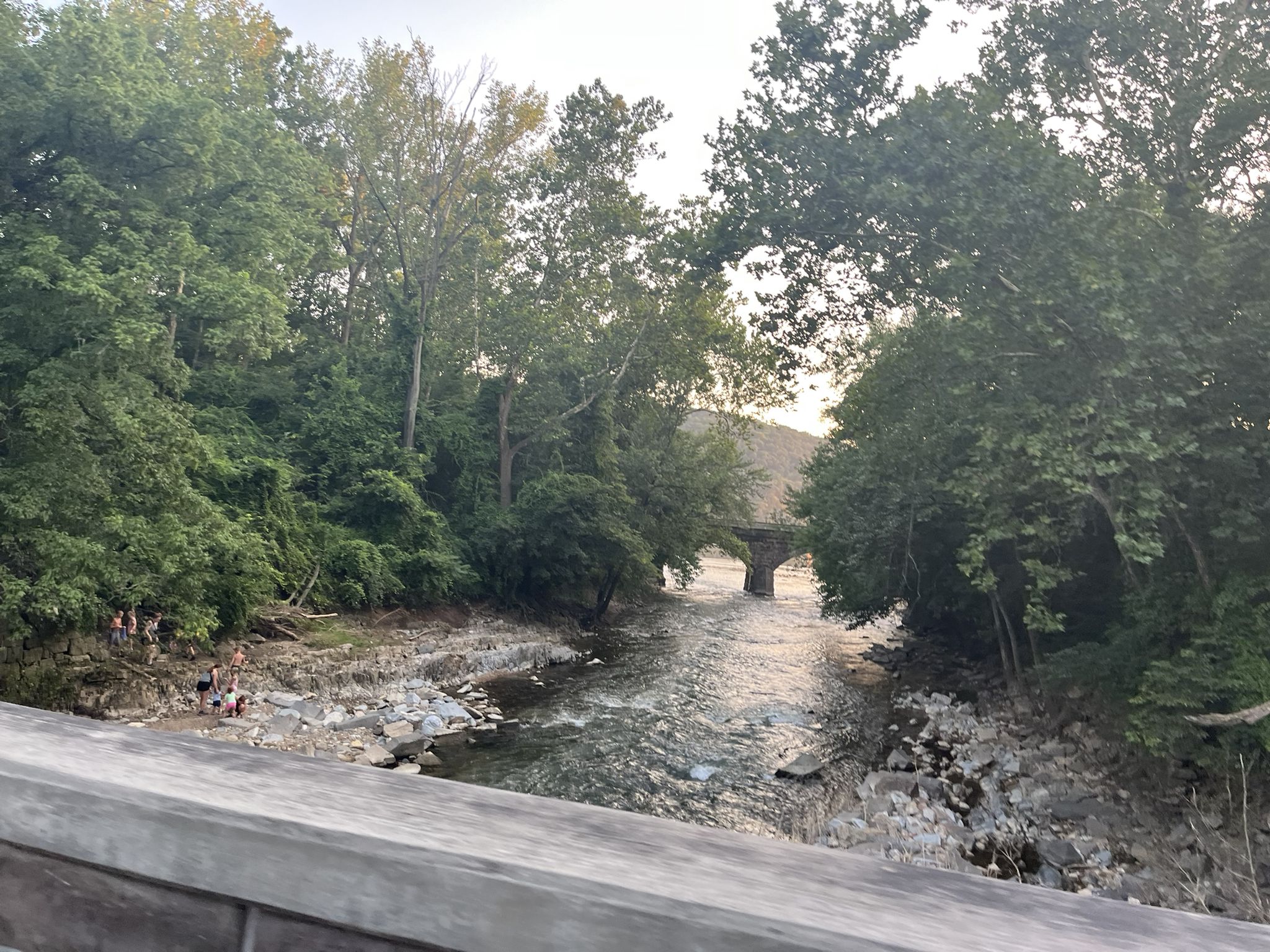 River flowing under a stone arch bridge, dense trees, people on riverbank at dusk. - Old Trolley Line Bridge loop from Marietta