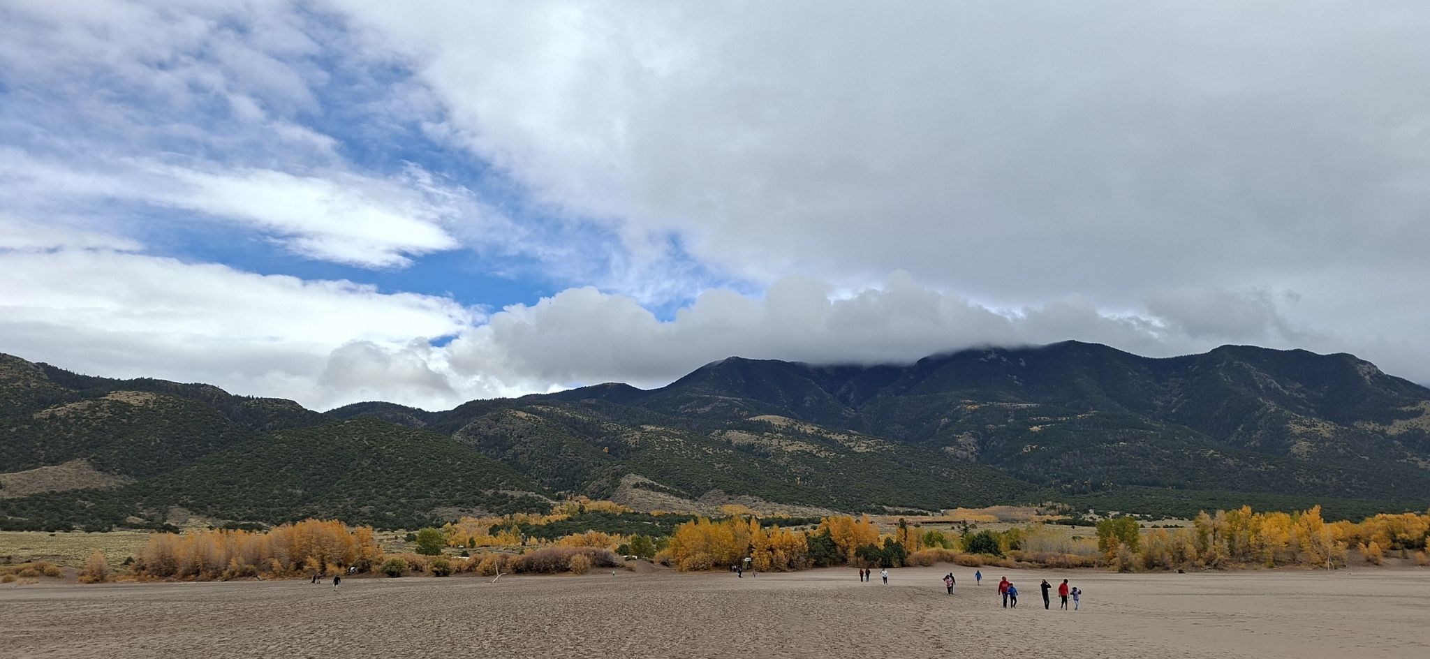 Groups of people walk across a wide sandy plain with mountains and autumn trees. - Sand Dunes – Medano Creek loop from High Dune