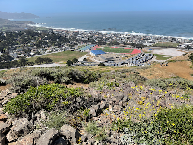 Elevated view of a sunny coastal town with buildings, fields, and ocean. - Milagra Ridge Loop