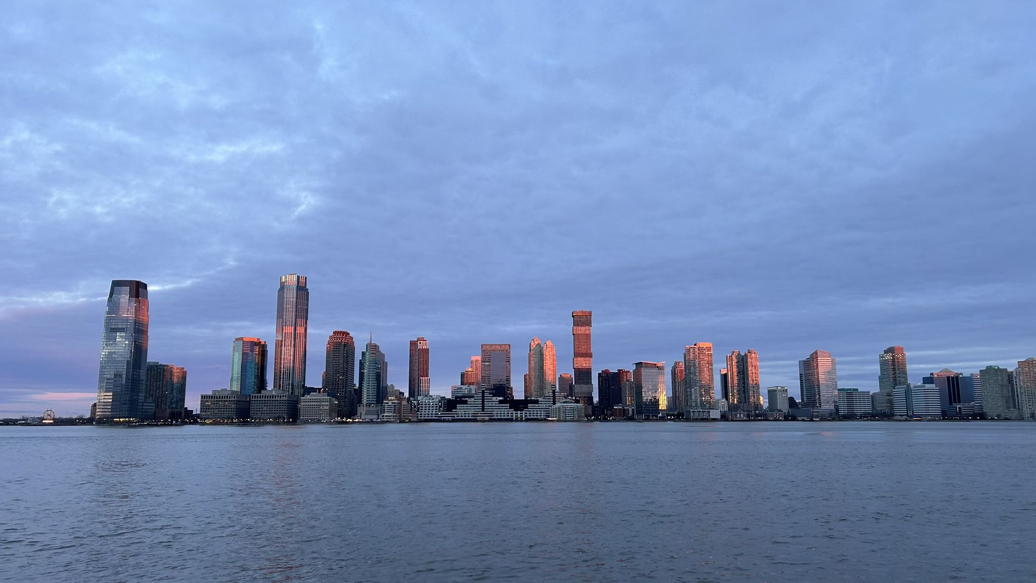 City skyline reflecting sunset colors on water under a cloudy sky. - Battery Park and Battery Park City Greenway Loop