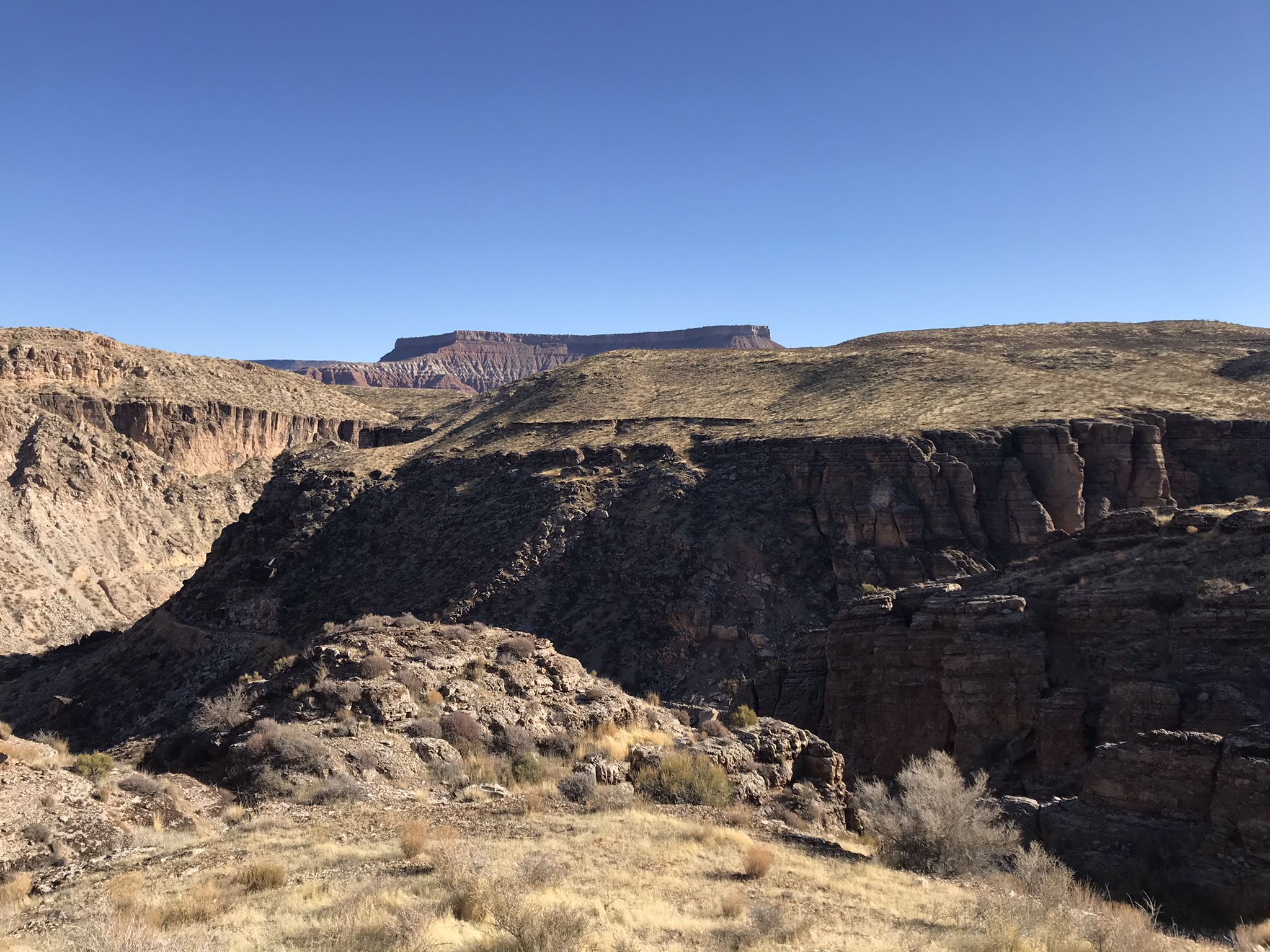 A wide desert canyon with layered rock formations under a clear blue sky. - Hurricane Rim Loop