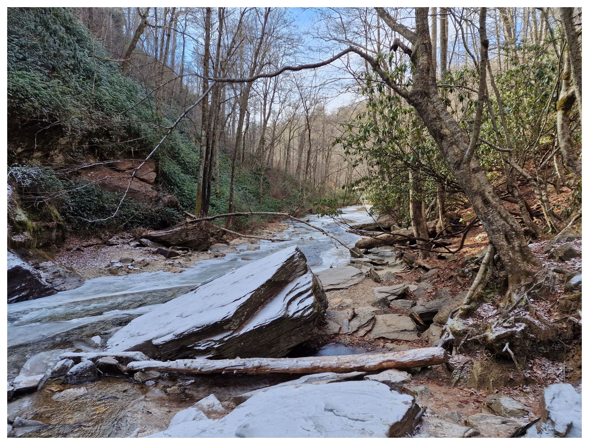 A snowy, rocky creek bed winds through a forest with bare trees. - Looking Glass Falls – Looking Glass Falls Viewpoint loop from Brevard