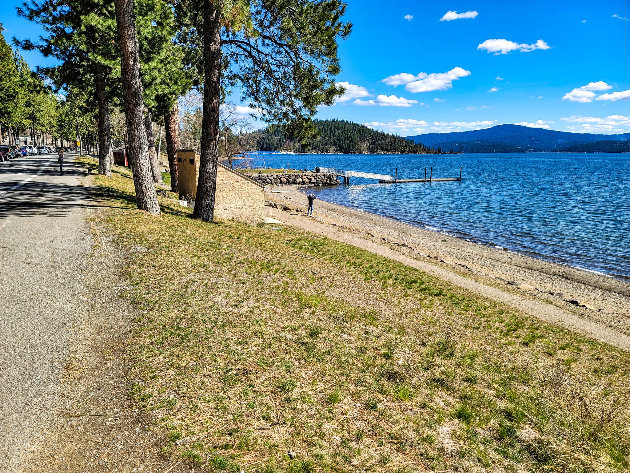 Paved path next to a sandy lake beach with a person and distant hills. - Hayden Lake loop