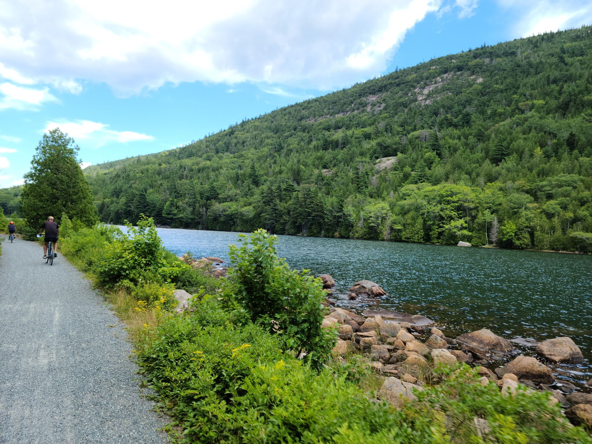 Person cycles on a gravel path beside a lake and forested hill. - Stone bridge – Bubble Pond loop from Parkman Mountain