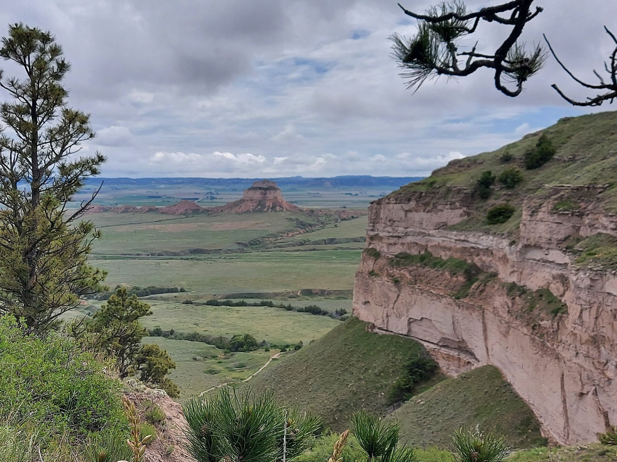 Green valley and steep cliffs framed by trees under a cloudy sky. - Scotts Bluff National Monument – Saddle Rock Trail loop from Scotts Bluff National Monument