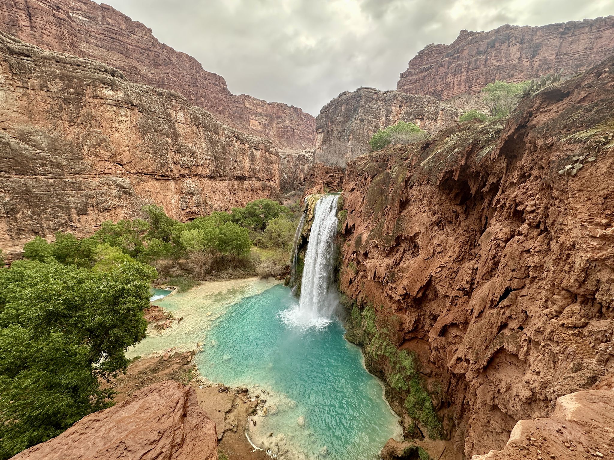 Waterfall plunging into a turquoise pool, surrounded by red rock cliffs under cloudy sky. - The Confluence from Supai Village