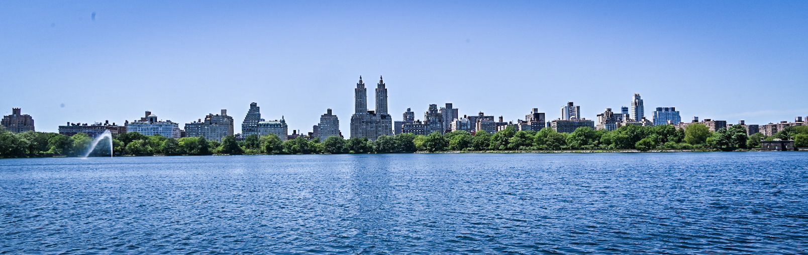 A wide blue lake with a city skyline and fountain under a clear sky. - Hudson River Greenway – Harlem Meer loop from 135th Street