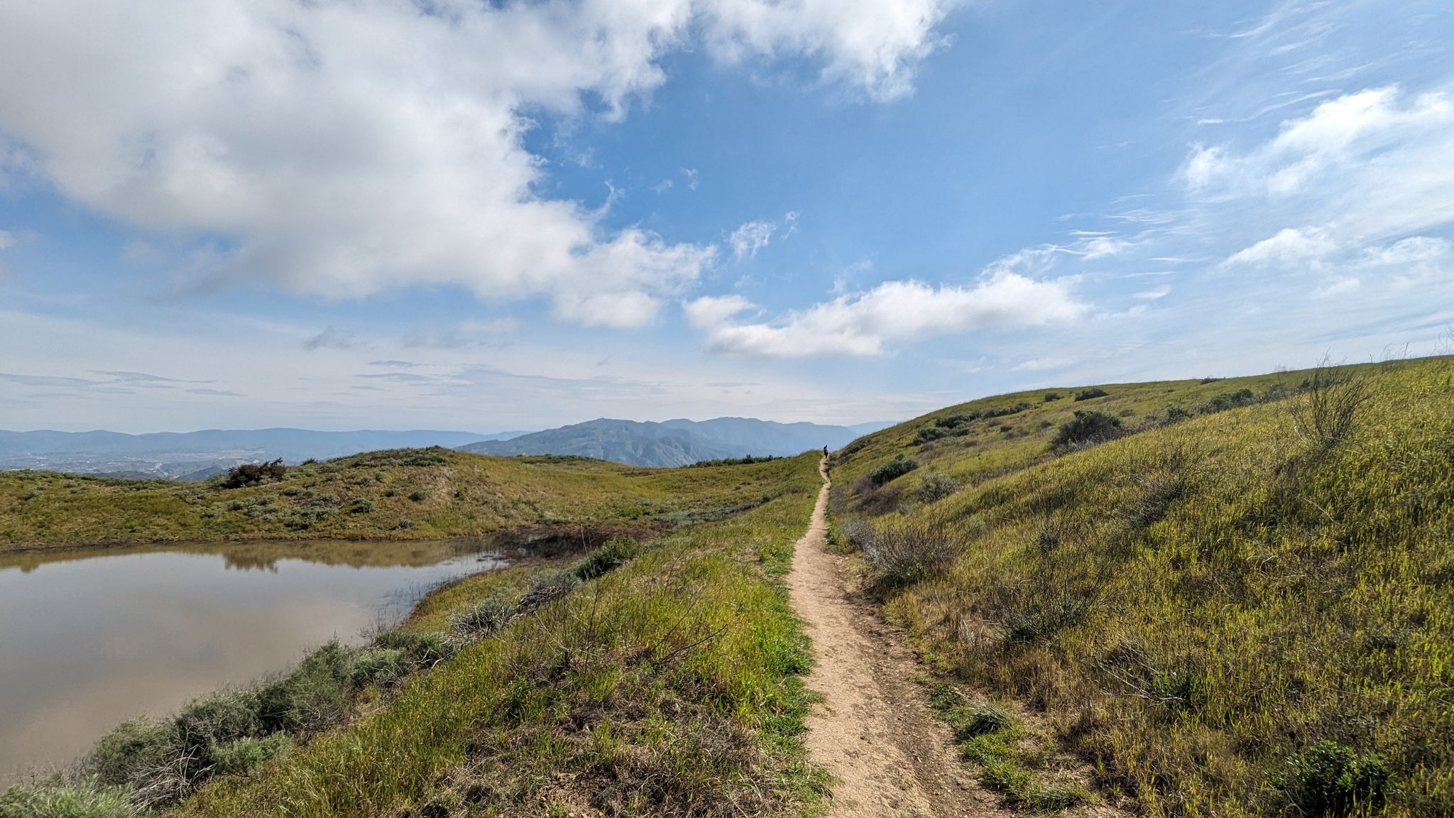 A dirt path alongside a calm pond, leading up a grassy hill. - O'Melveny Park and Mission Point Loop