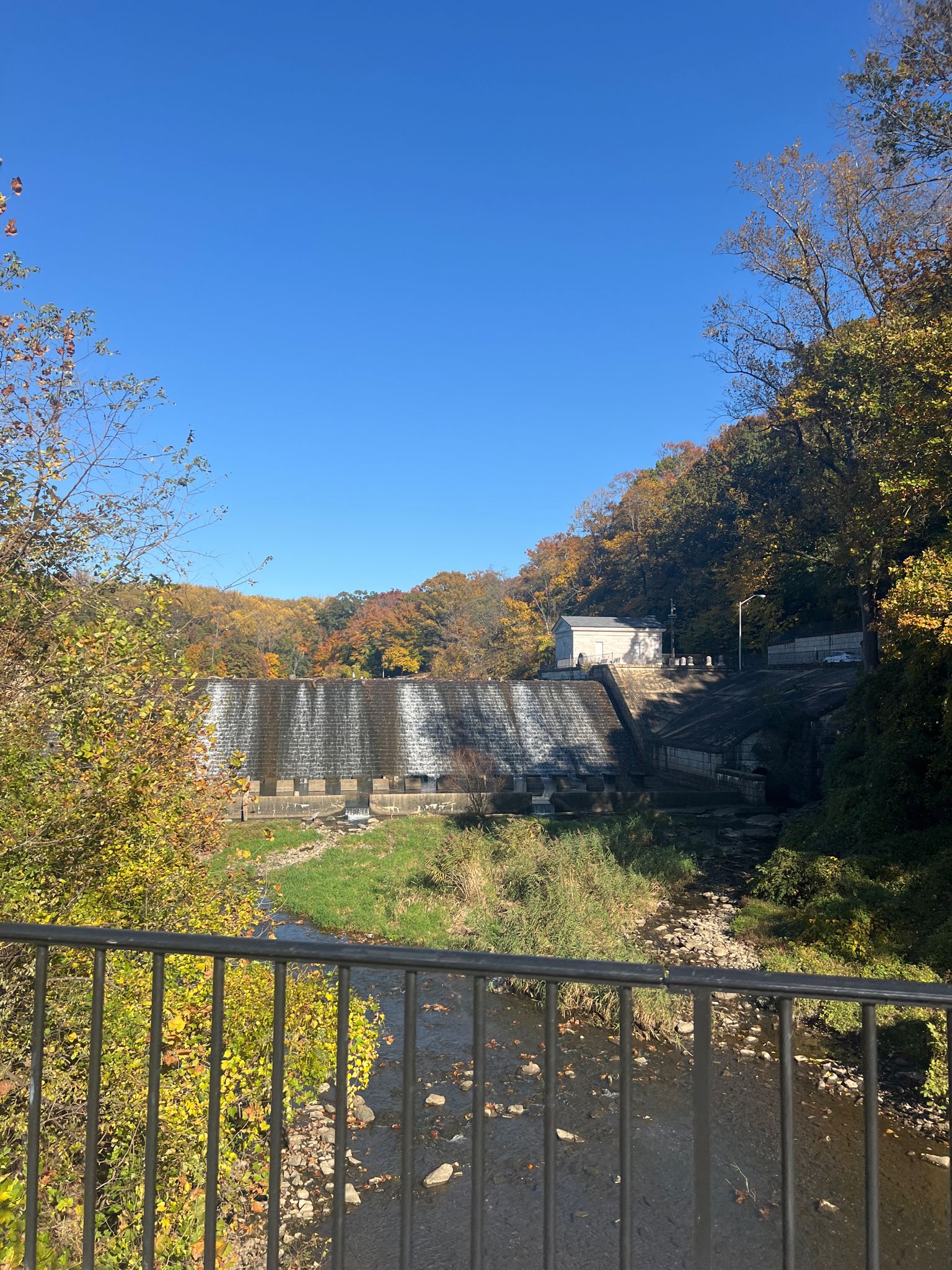 Dam with water cascading over it, surrounded by autumn trees under clear sky. - Lake Roland Red Trail Loop