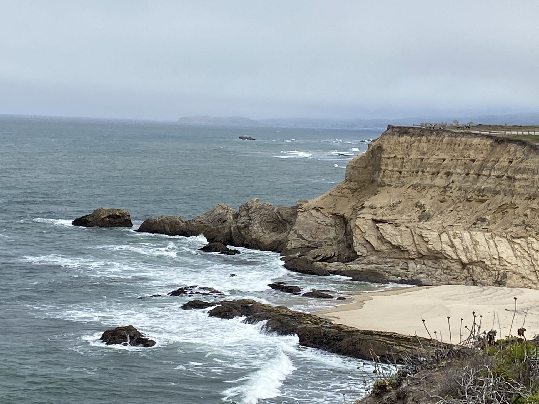 Overcast view of rugged cliffs, a small sandy beach, and crashing ocean waves. - Half Moon Bay State Beach loop from Johnston Ranch