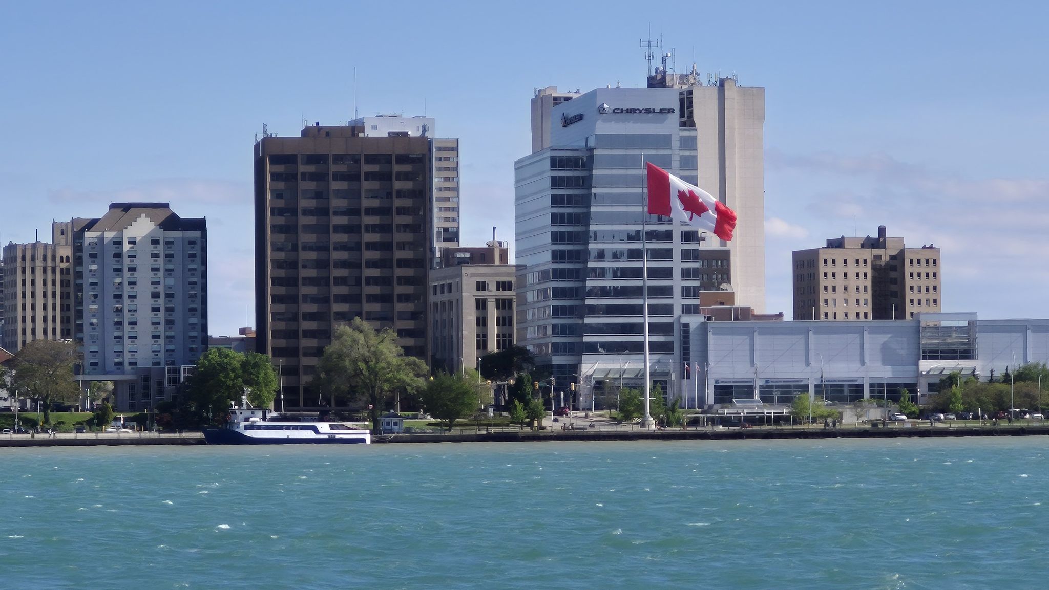 A city skyline with tall buildings, a Canadian flag, and a boat on blue water. - Woodward Avenue – Campus Martius loop from Huntington Place